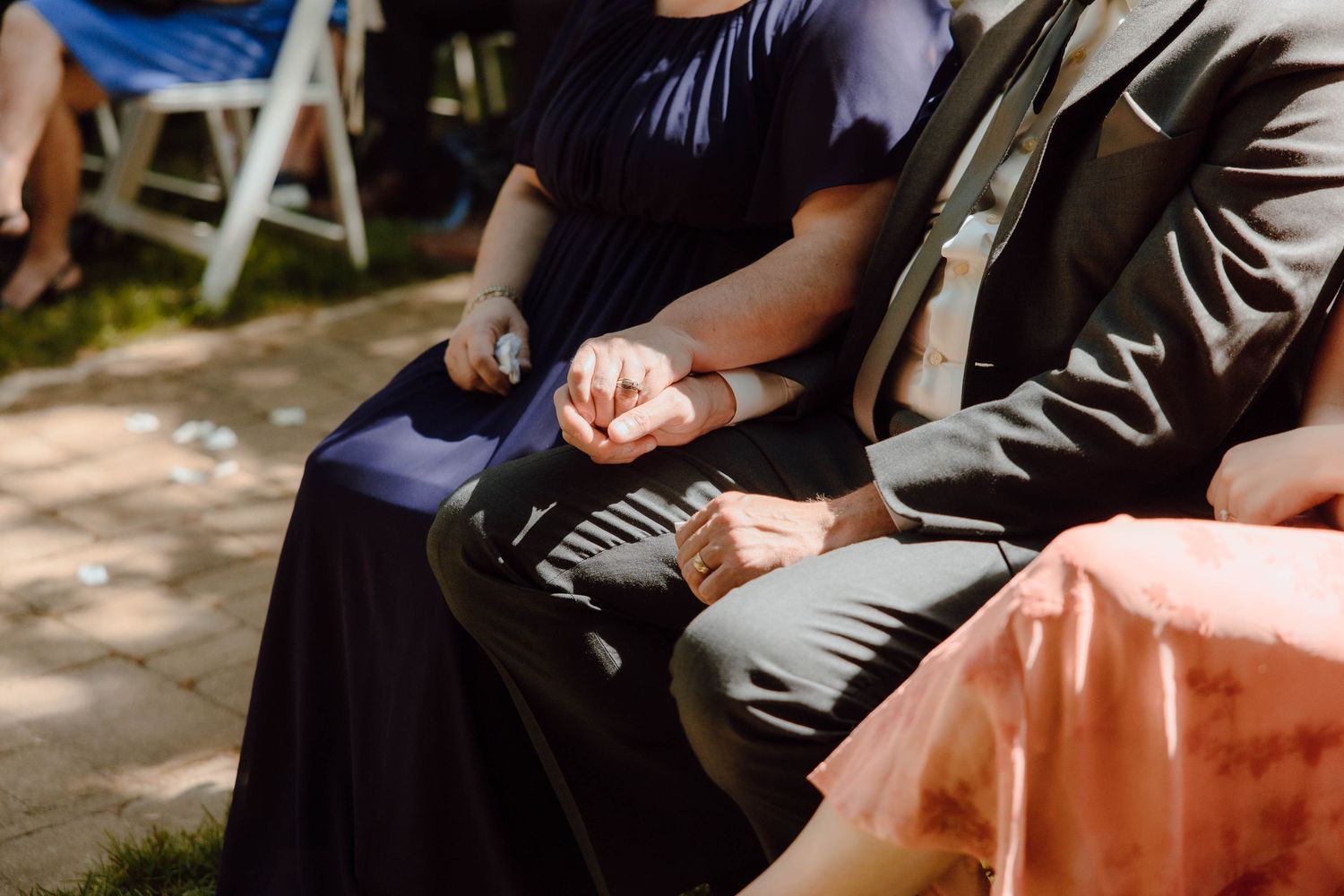 Wedding guests in formal attire hold hands tenderly while seated during an outdoor ceremony in sunlight.