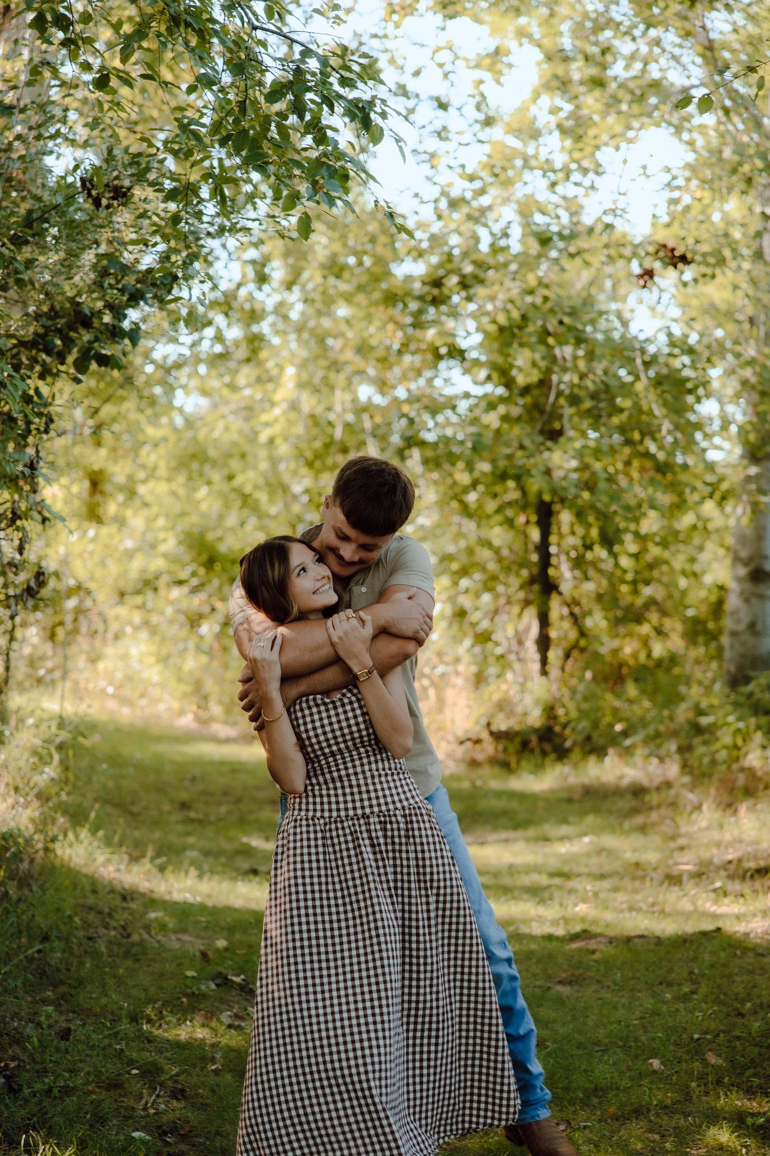A couple embraces tenderly in a sunlit forest path, woman in checkered dress, man hugging her from behind.