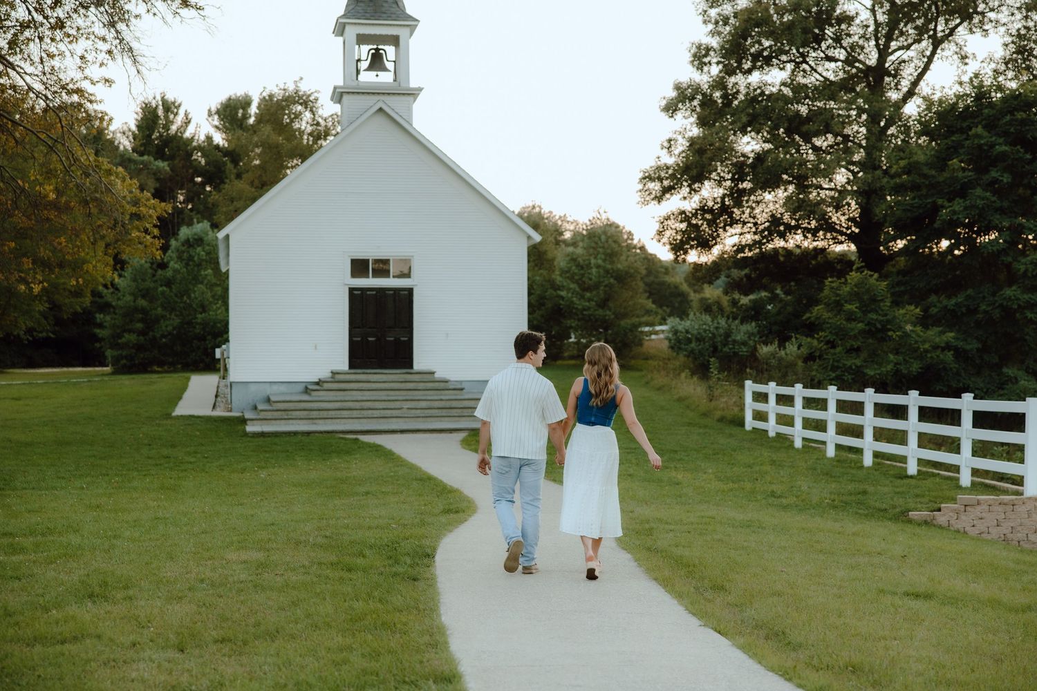 Couple holding hands walking toward a white country church on a curved path surrounded by green grass and trees.