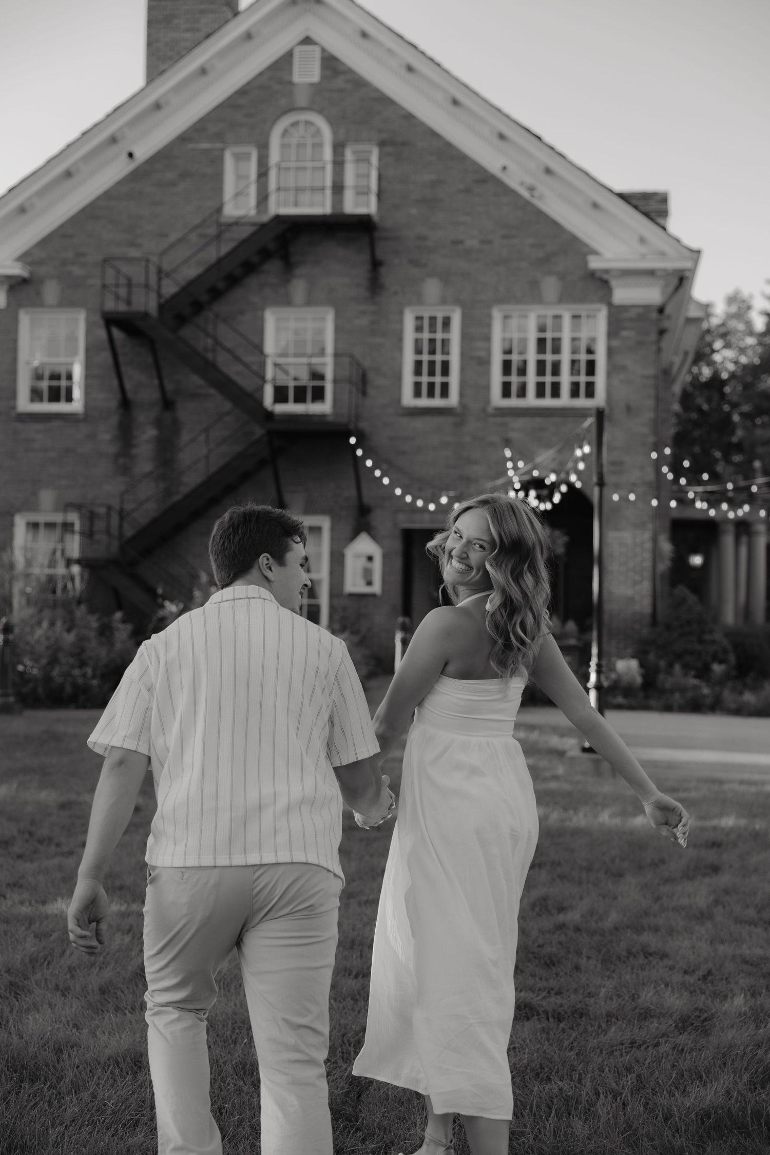 Black and white photo of a couple holding hands walking toward a brick building with string lights at dusk.