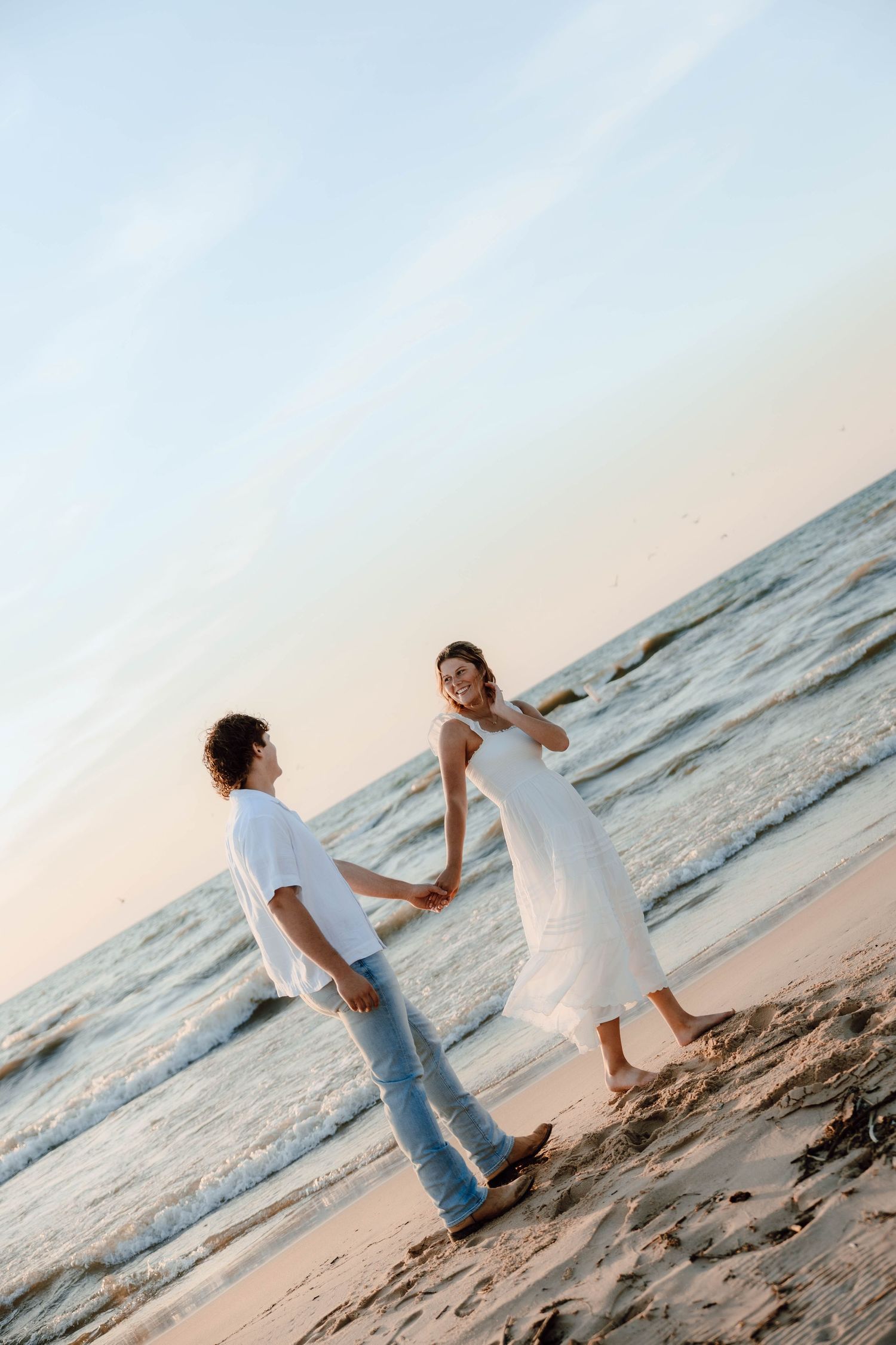 Romantic couple holding hands on a sandy beach at sunset, woman in white dress, man in jeans and white shirt.