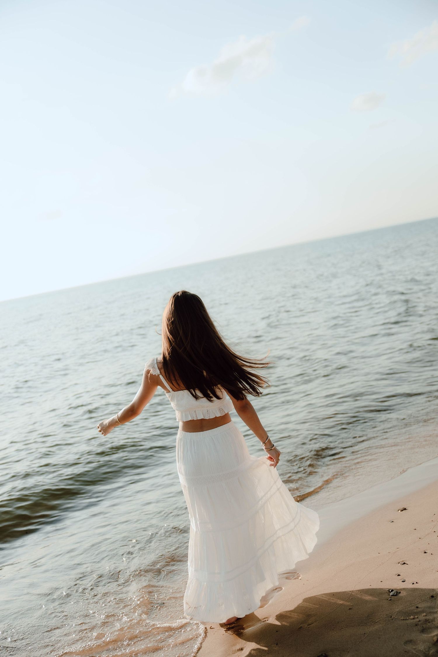 Woman in white crop top and flowing skirt walking barefoot on sandy beach with ocean waves.