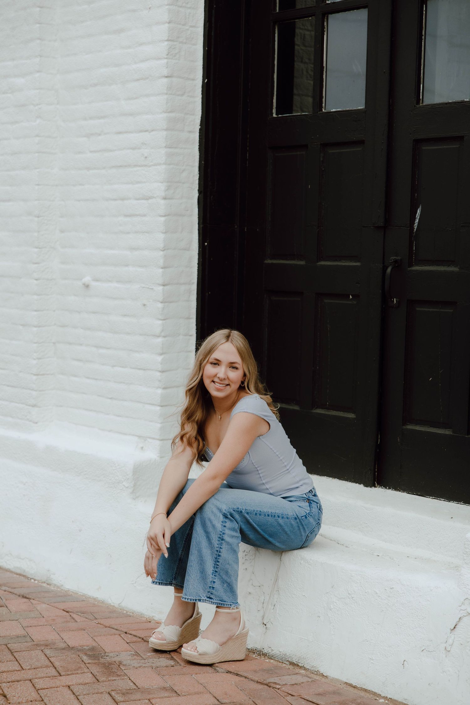 Young woman in jeans and gray top sitting on white brick ledge near black door, smiling at camera.
