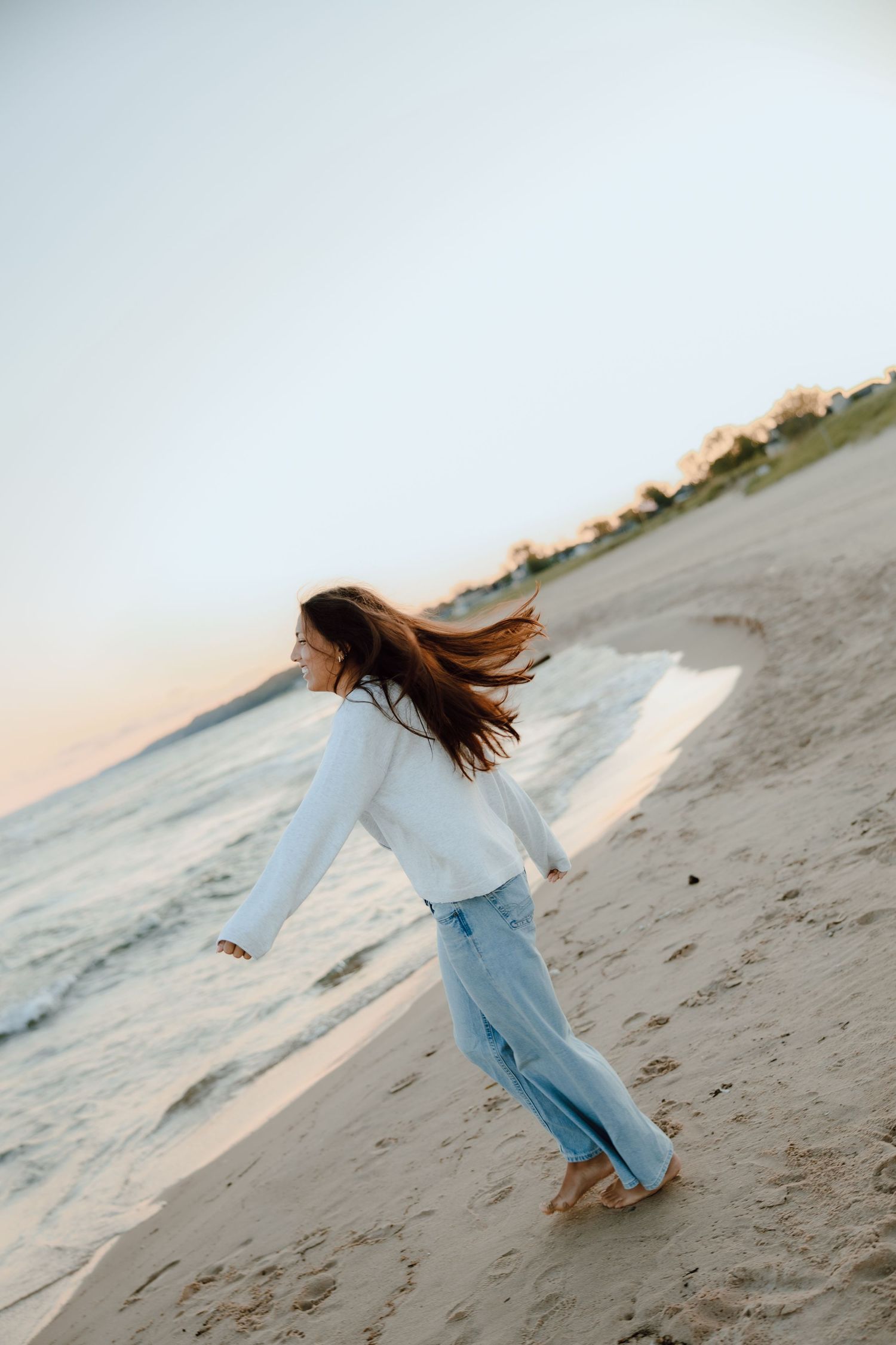 A young woman in a white top and jeans runs barefoot on a sandy beach at sunset with her hair blowing in the wind.
