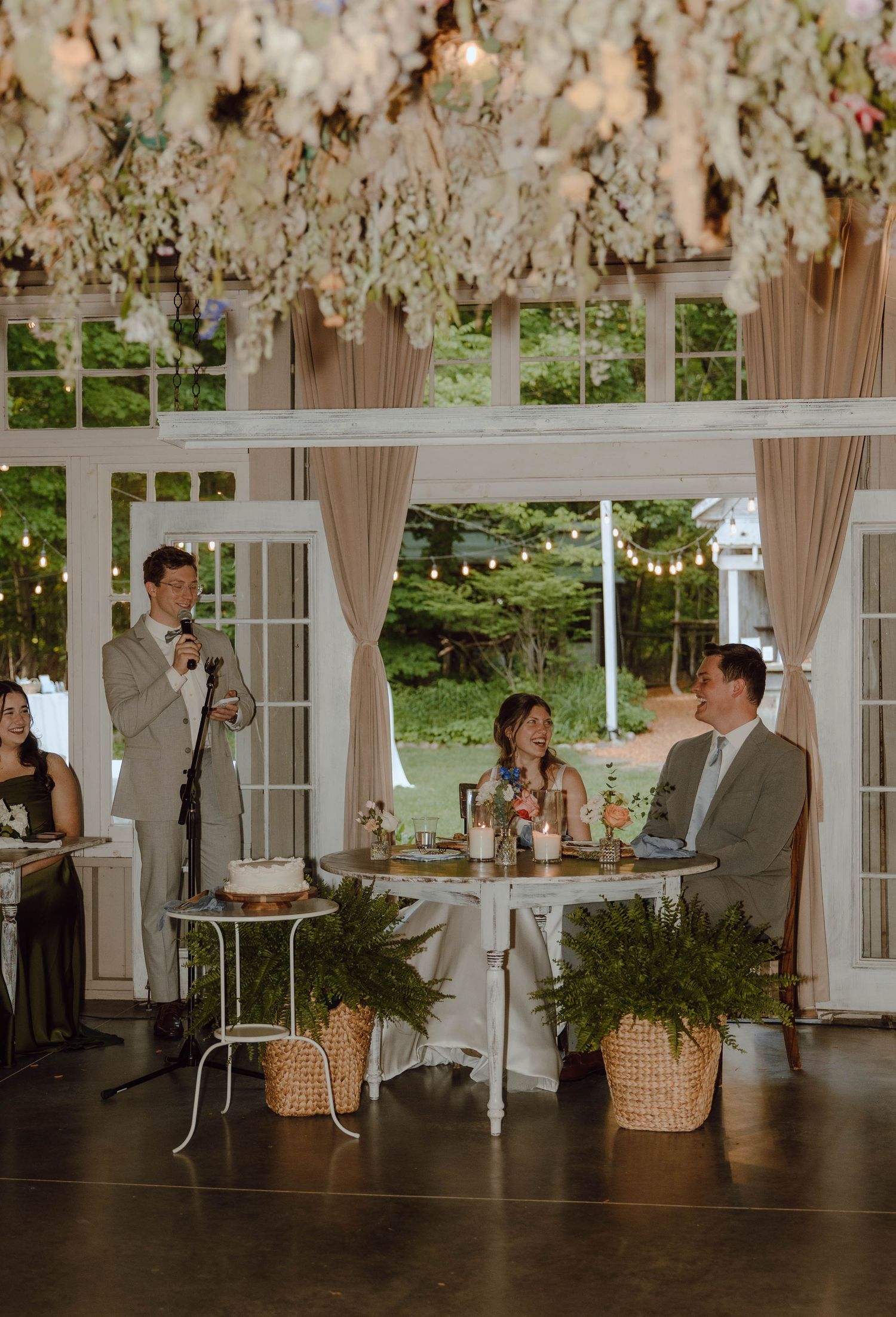 Groom delivers a heartfelt toast to the bride and groom seated at a decorated reception table indoors.
