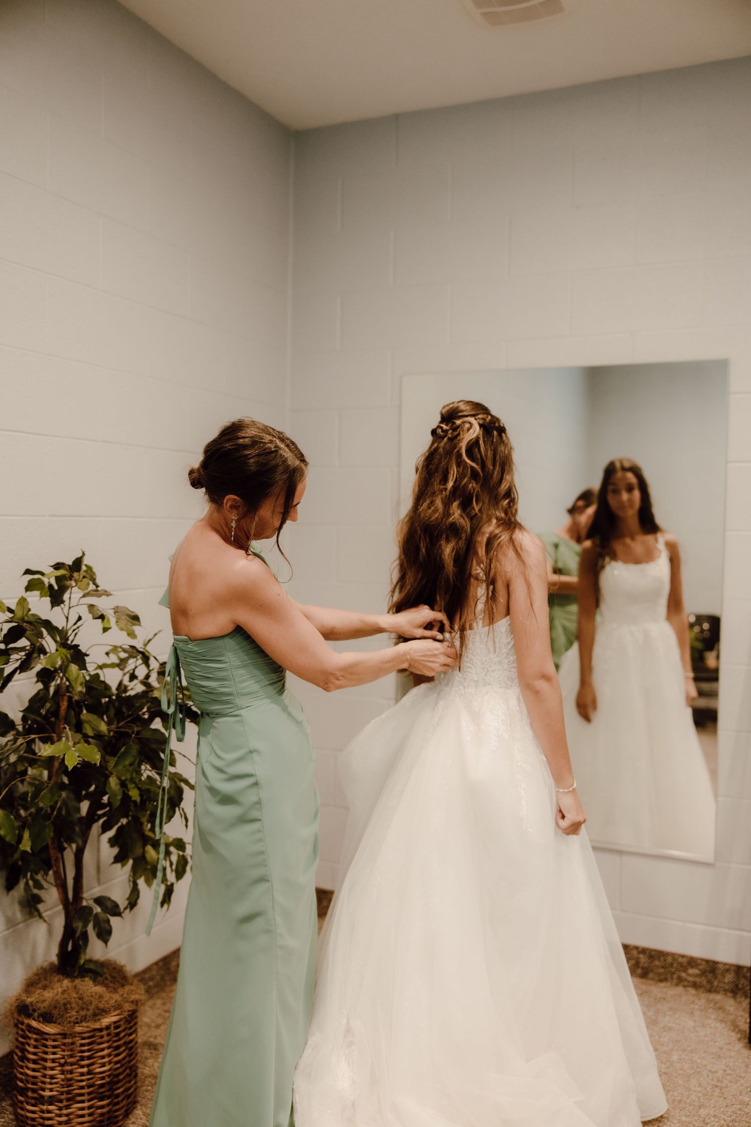 Bridesmaid in sage green dress helping bride with wedding gown while reflected in mirror.