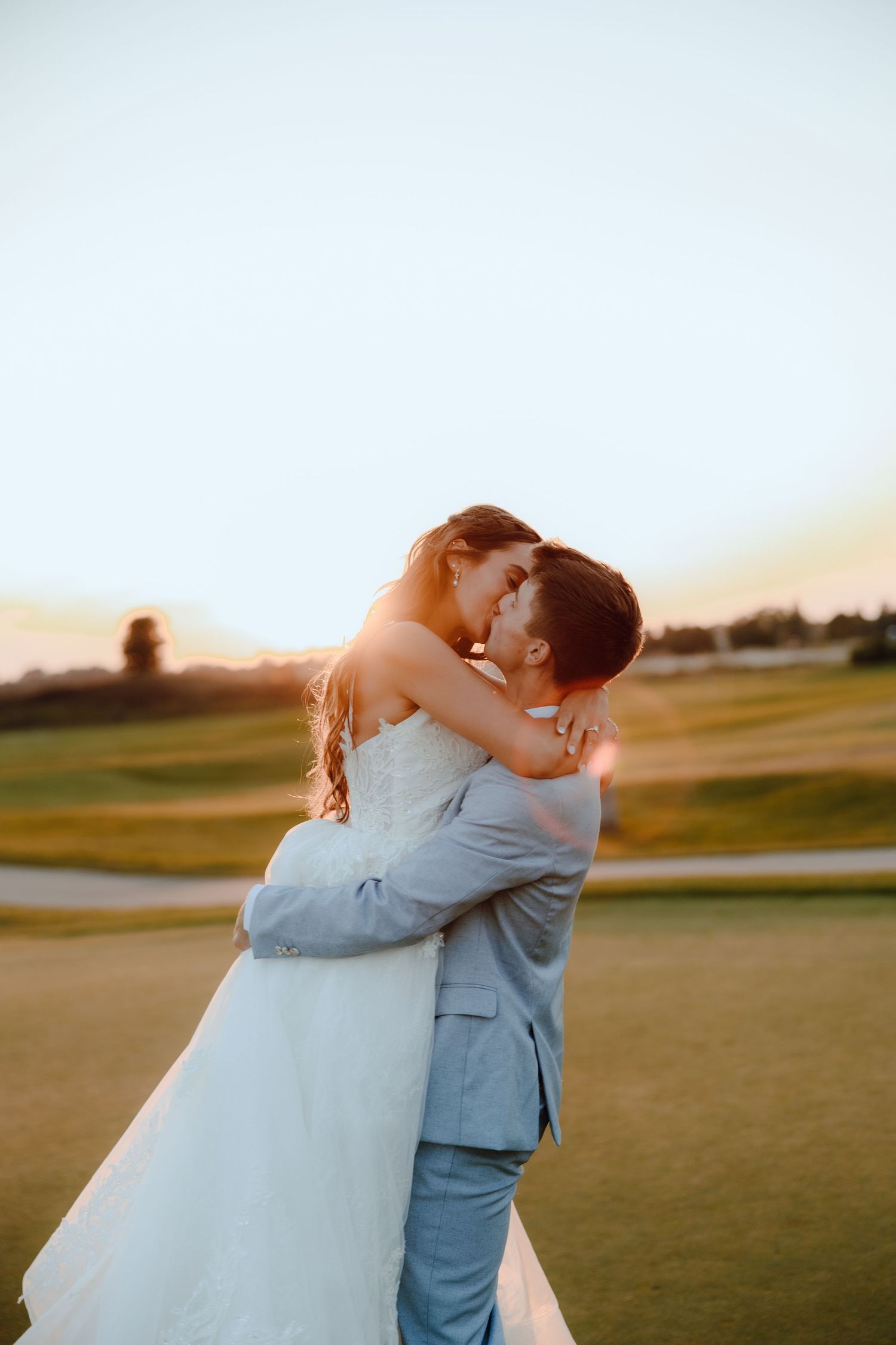 Bride and groom embrace at sunset on a golf course, sharing a romantic moment in wedding attire.