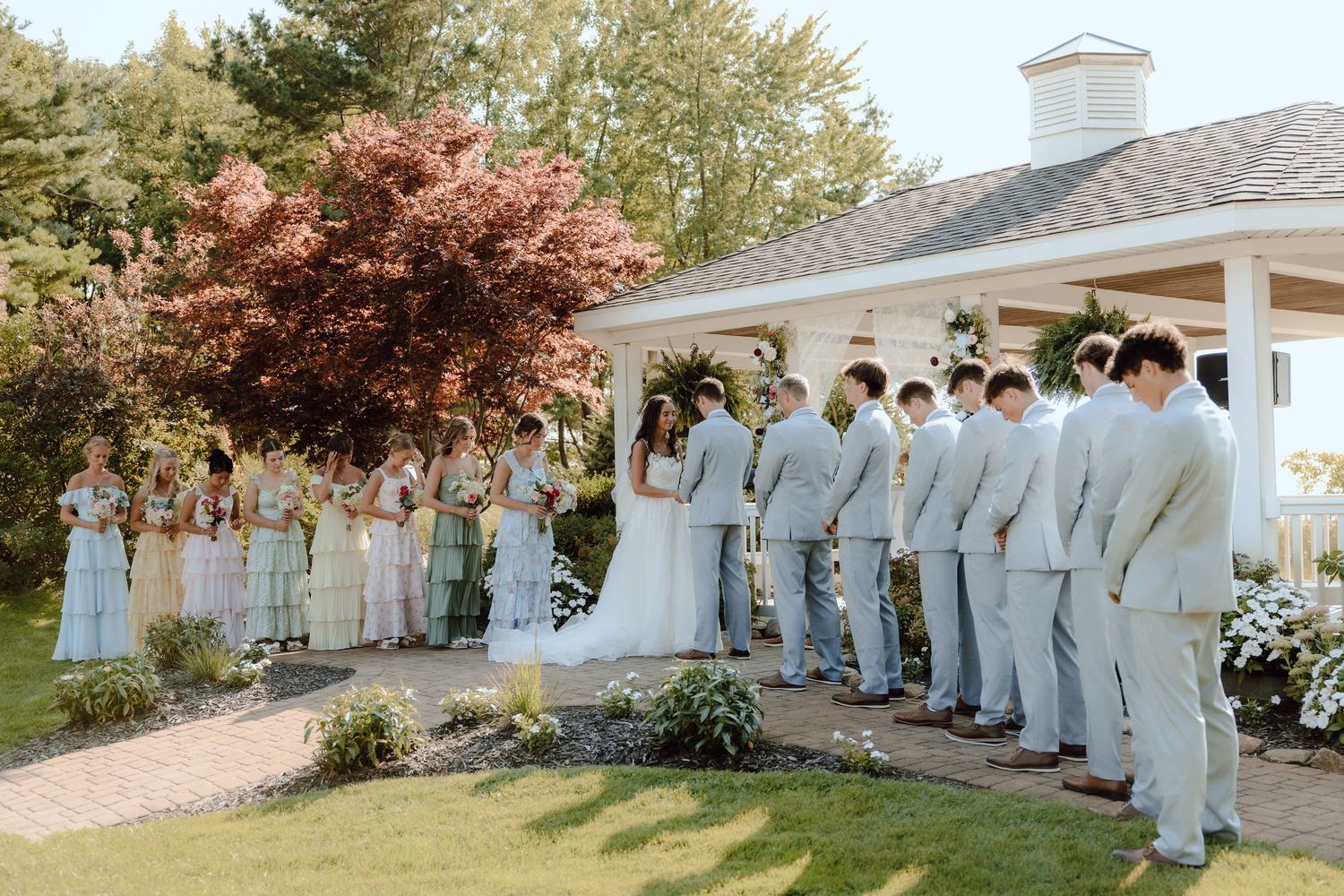 Full wedding party gathered outside white gazebo venue with lush greenery and red maple tree.