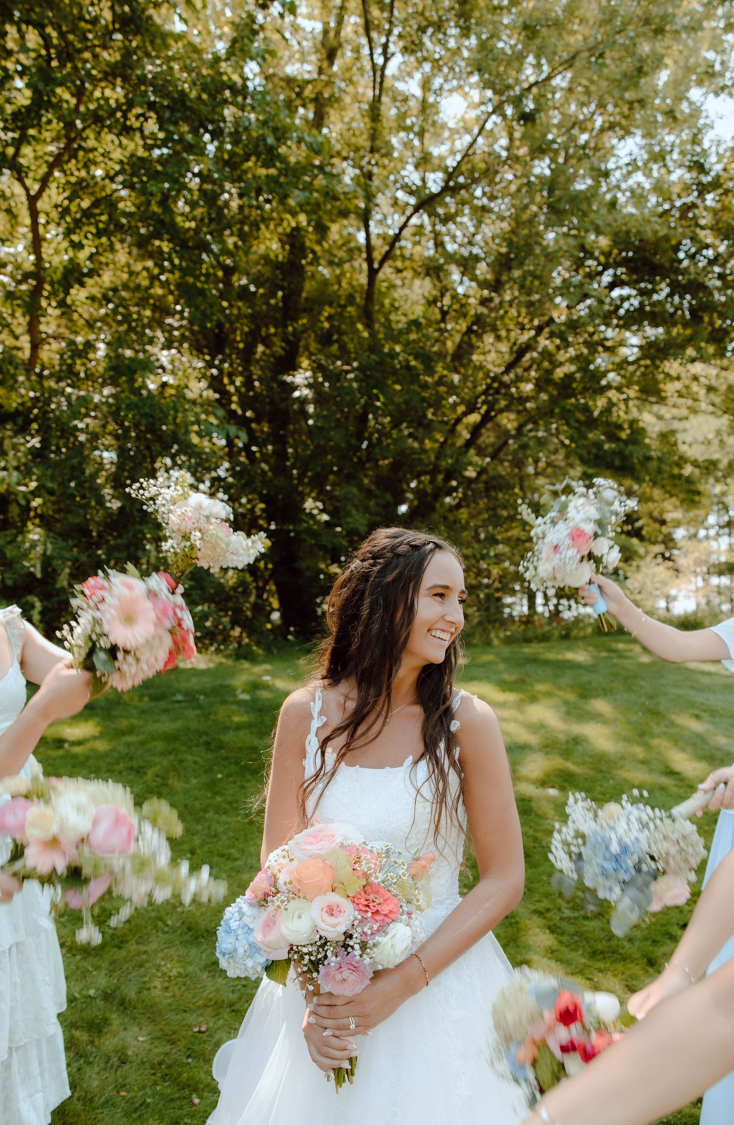 Smiling bride in white gown holds colorful bouquet surrounded by bridesmaids with flowers outdoors.