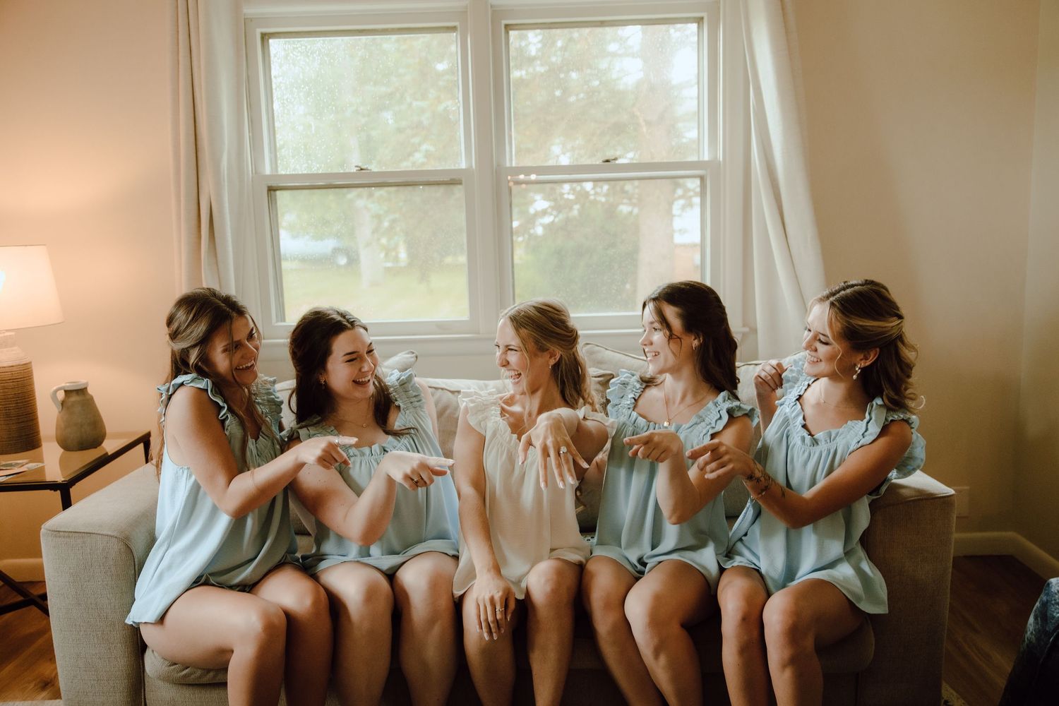 Five bridesmaids in light blue robes laughing together on a couch near a bright window before a wedding.