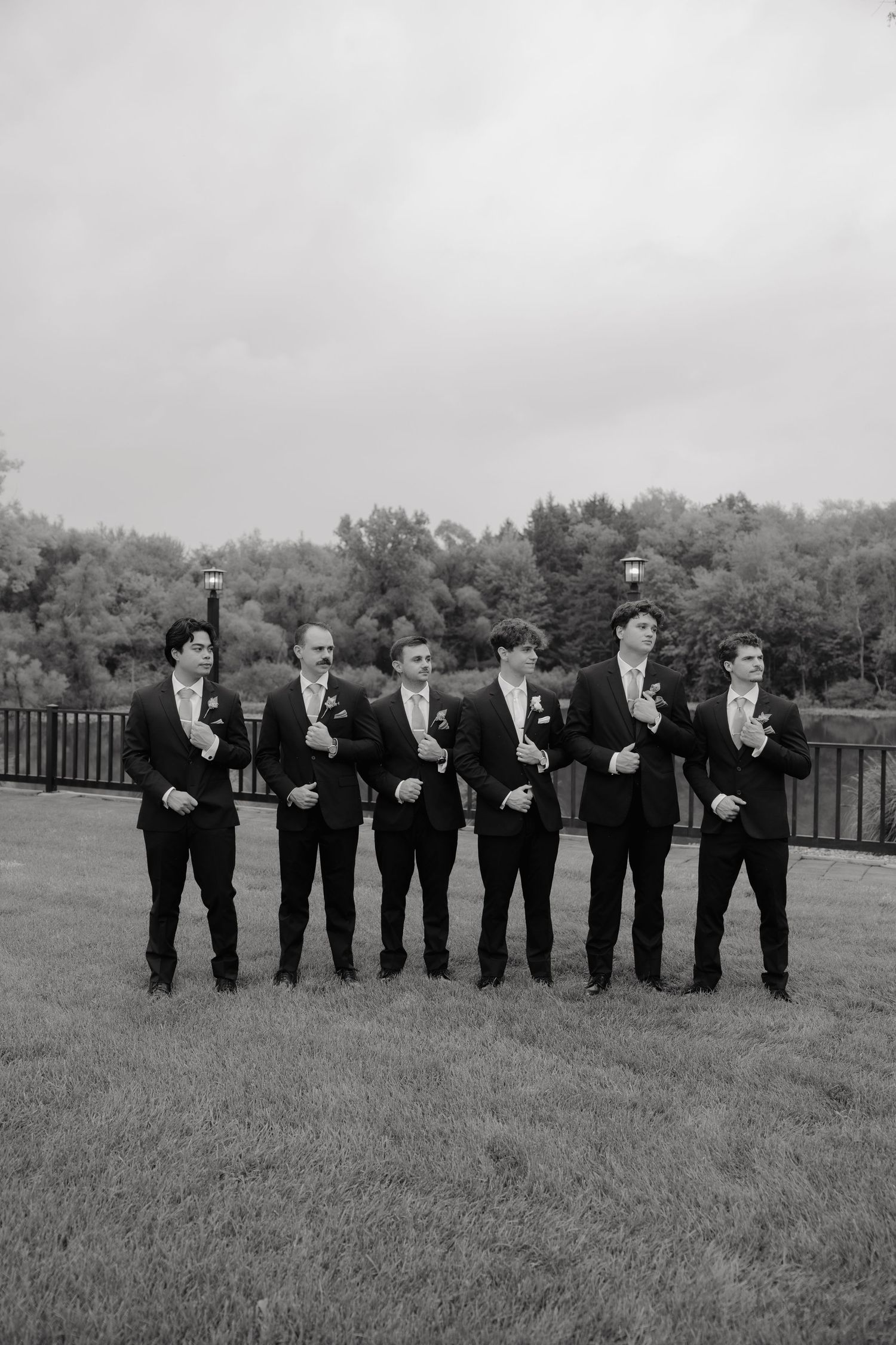 Black and white photo of six groomsmen in suits posing on lawn outdoors.