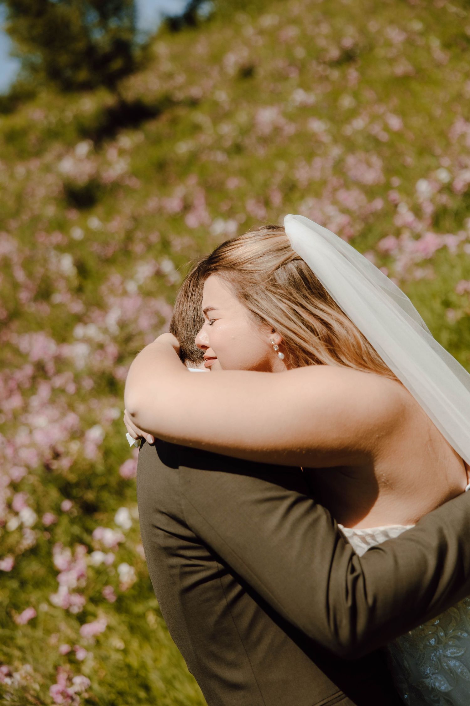 Bride in veil embraces groom in green suit in a sunlit field of pink wildflowers.
