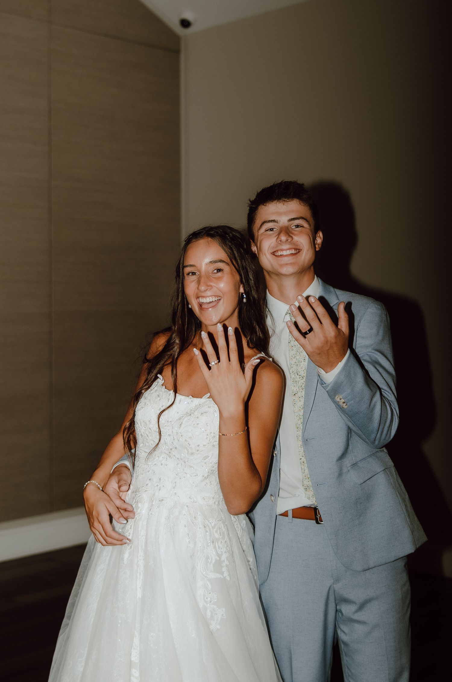 Newlyweds in wedding attire smiling and showing off wedding rings, bride in white gown and groom in light blue suit.