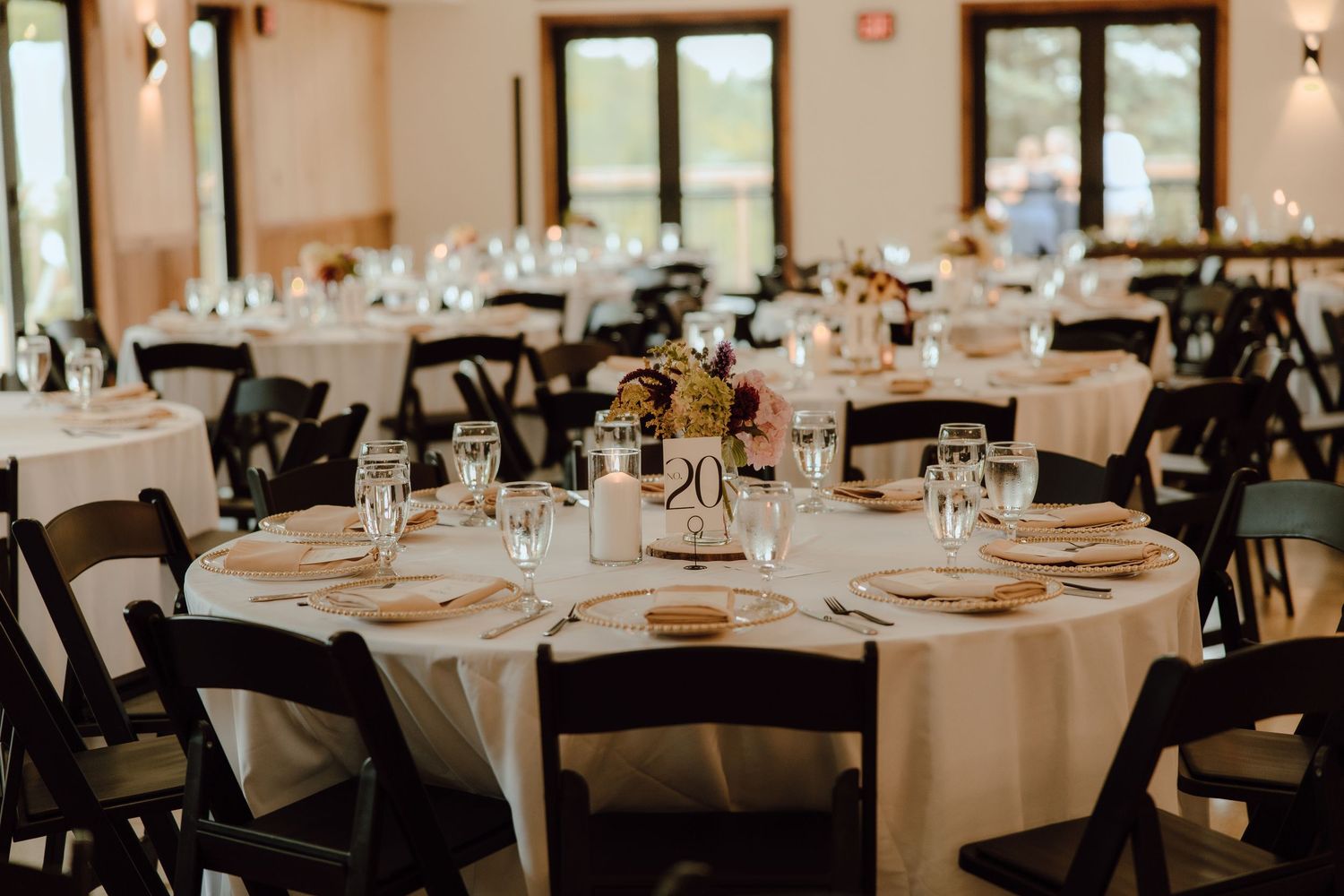 Elegant wedding reception venue with round tables, white linens, black folding chairs, and floral centerpieces in a bright hall.