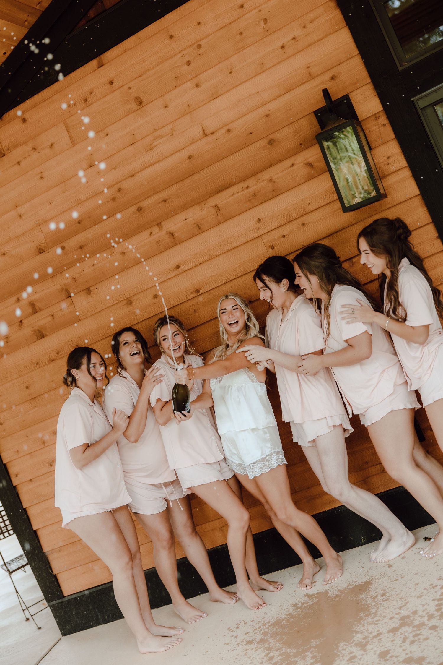 Bride and bridesmaids in matching pink pajamas celebrate with champagne outside a rustic wooden cabin.