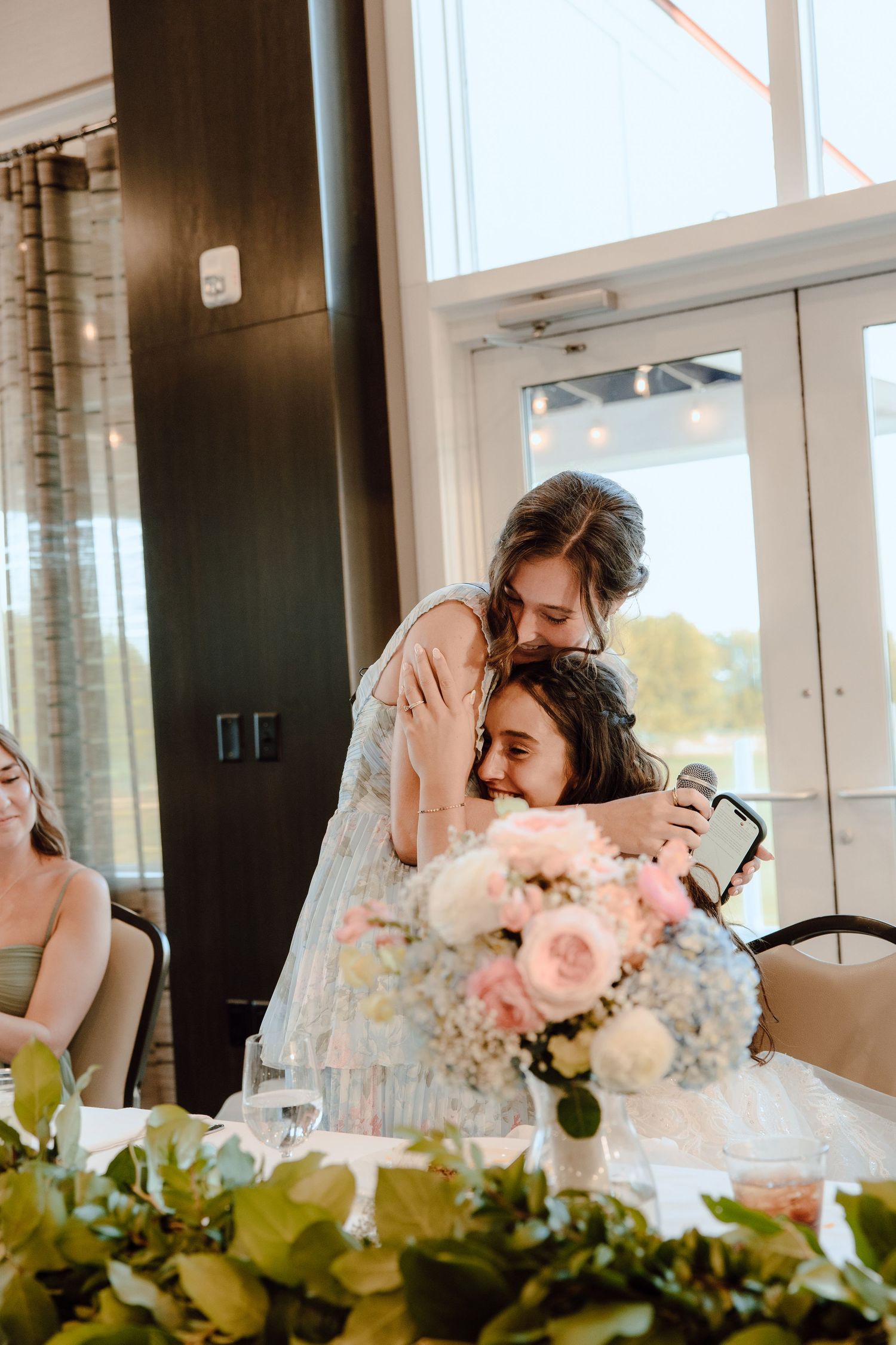 Bridesmaid embraces seated bride during heartfelt wedding reception speech at decorated head table.