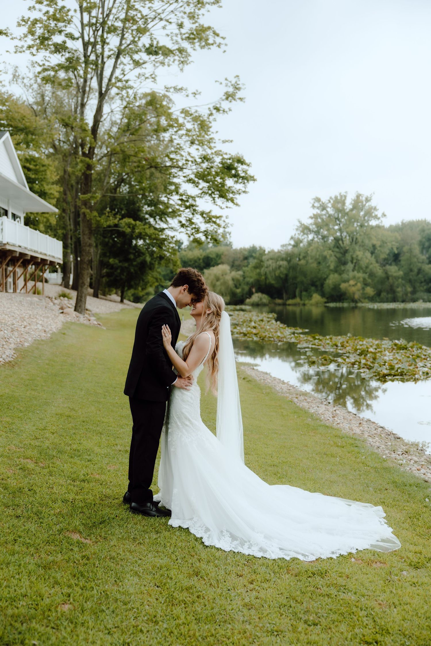 Bride and groom share a kiss on green lawn beside a serene lily pad pond on wedding day.