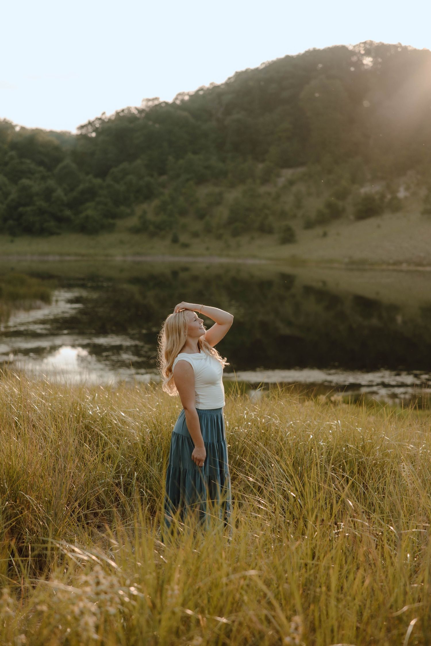 Blonde woman in white top and blue skirt standing in tall grass near a reflective lake at sunset.