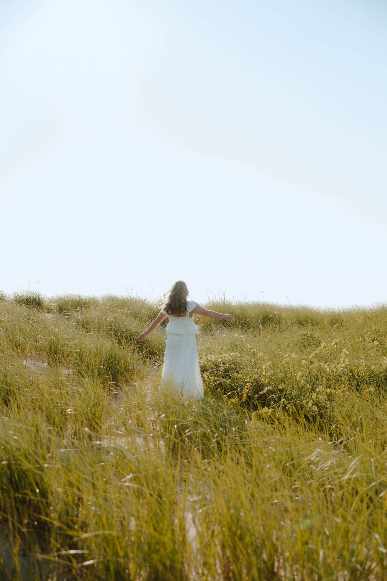 Woman in white dress standing in tall green grass field with arms outstretched, viewed from behind on a sunny day.