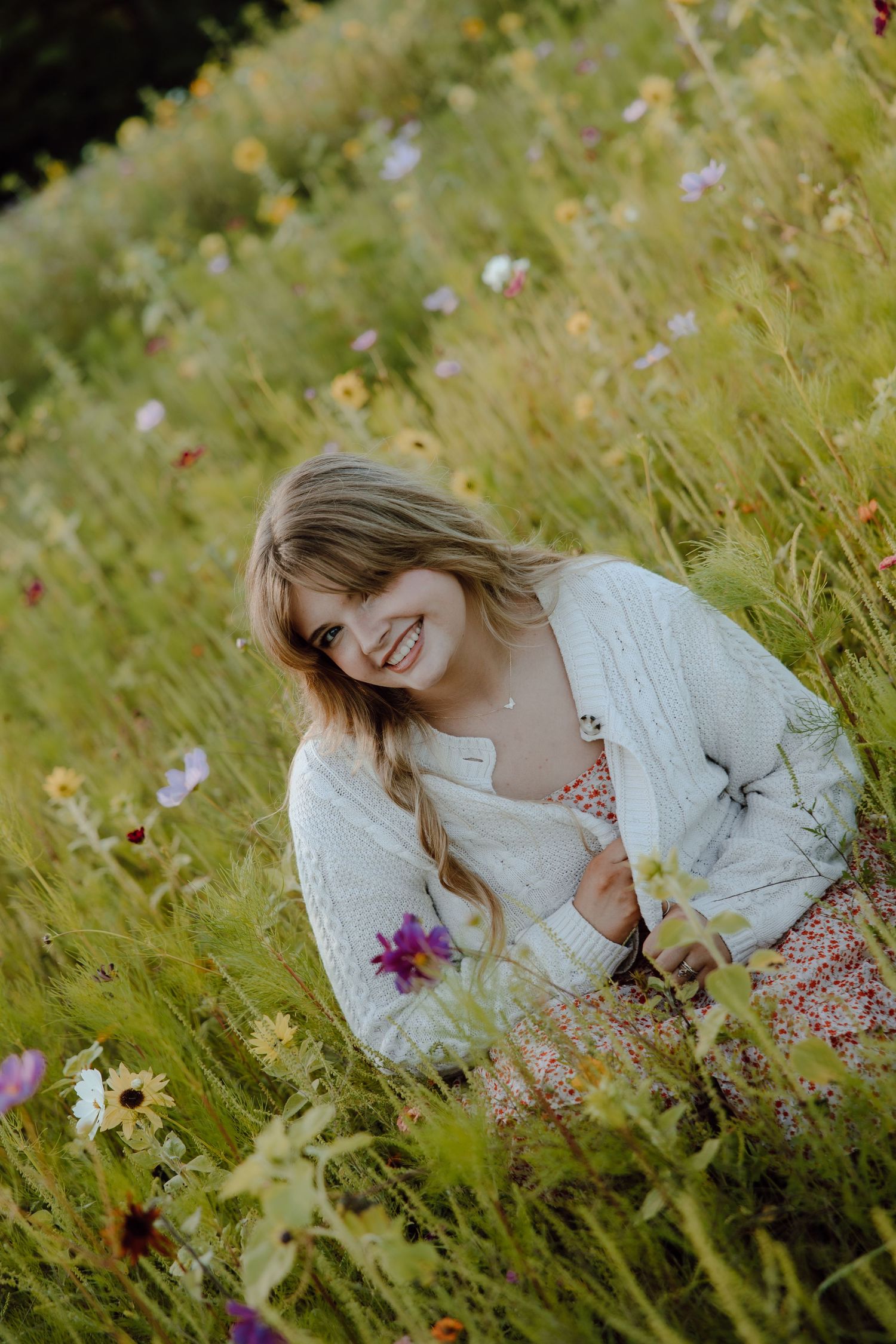 Smiling young woman in white cardigan sitting among colorful wildflowers in a lush green meadow.