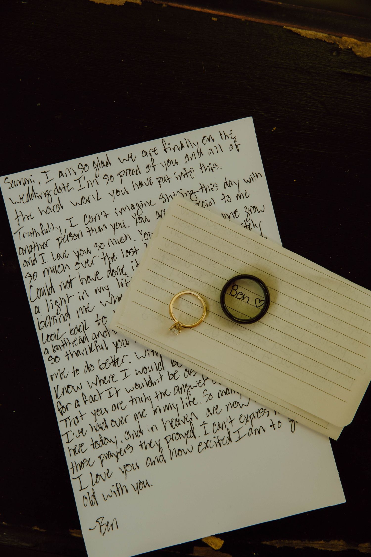 Handwritten wedding letter on lined paper with gold engagement ring and dark wedding band on wooden surface.