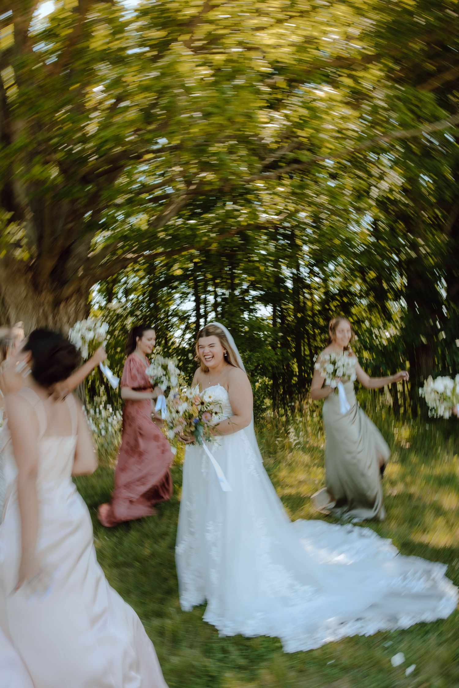 Bride in white gown laughs with bridesmaids in colorful dresses outdoors, surrounded by lush green trees.