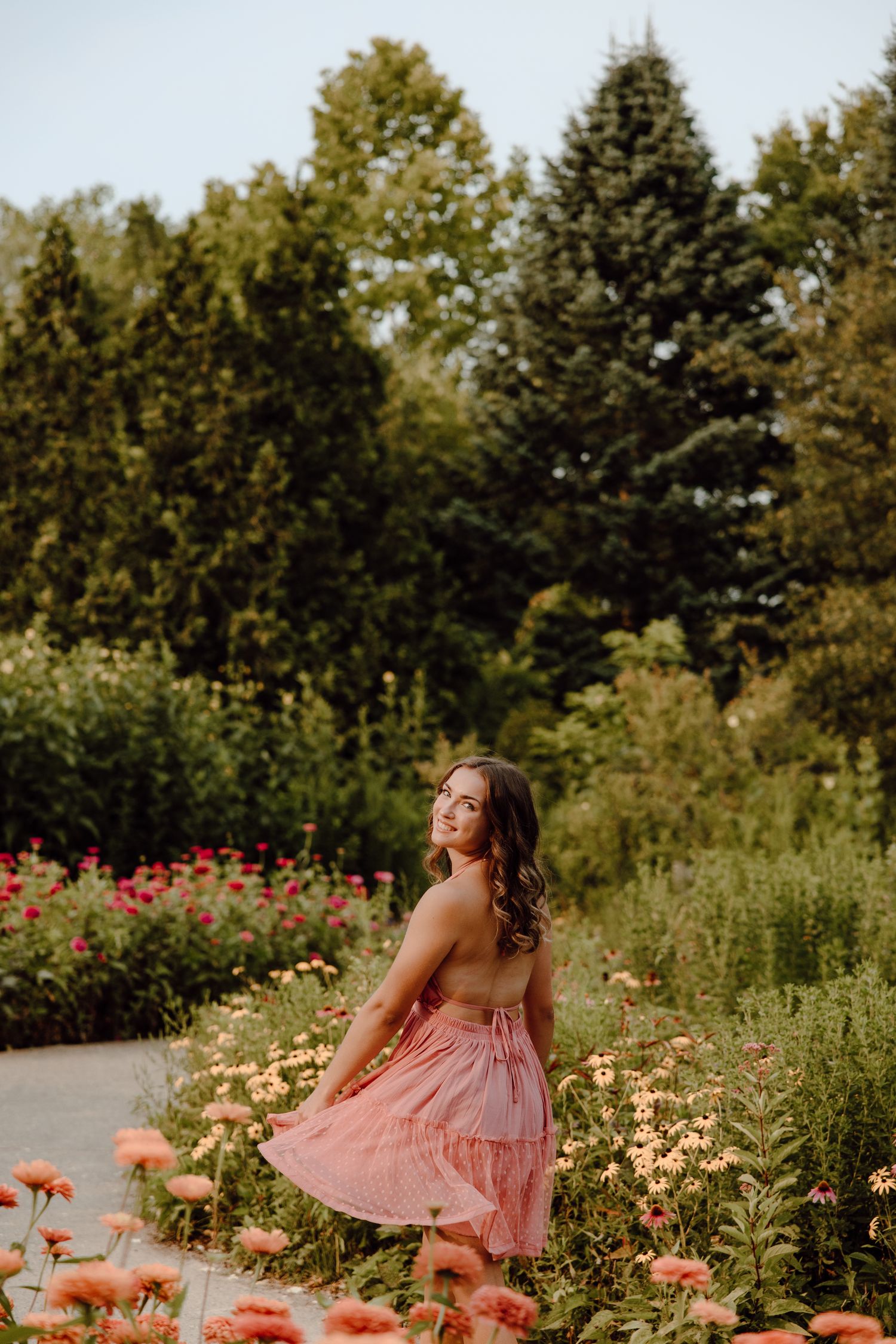 A person in a pink dress poses gracefully among blooming coral flowers in a garden setting.