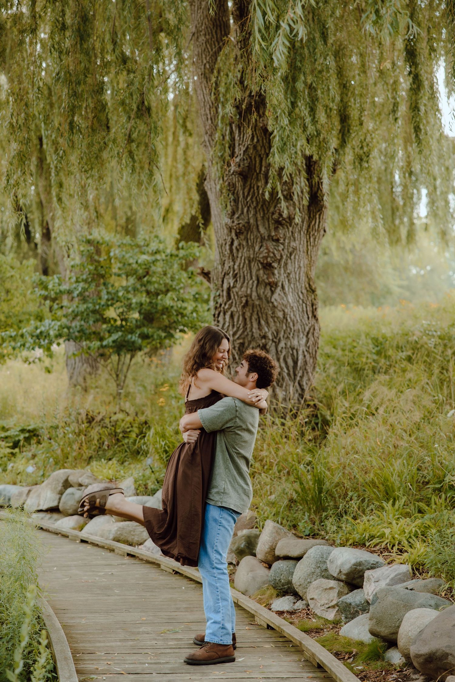 Couple embracing on a wooden boardwalk under a willow tree, surrounded by lush greenery and rocks.