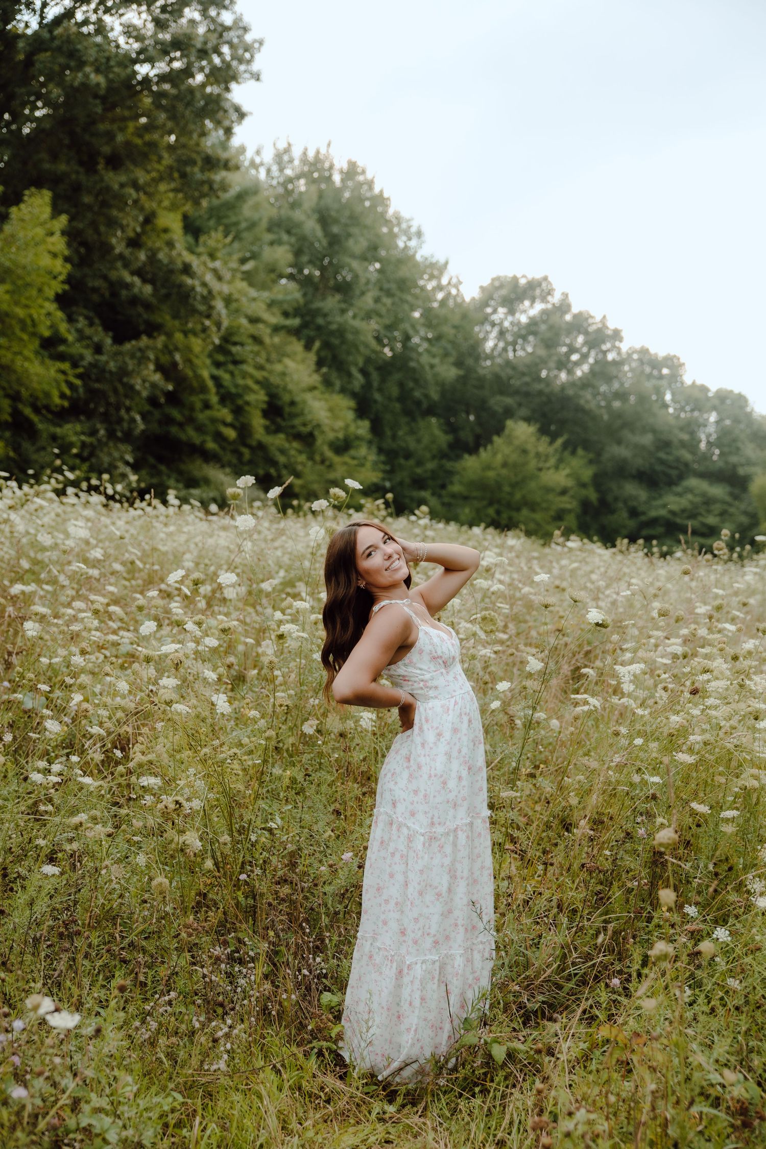 Young woman in floral maxi dress posing in wildflower meadow with lush green trees in background.