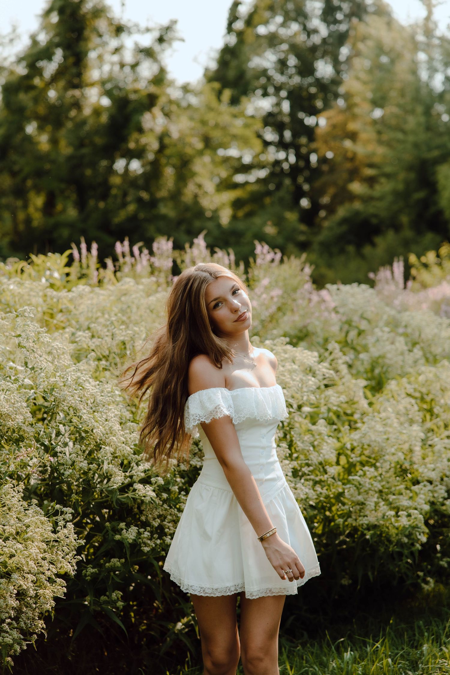 Young woman in white off-shoulder mini dress standing among wildflowers in a lush green garden setting.