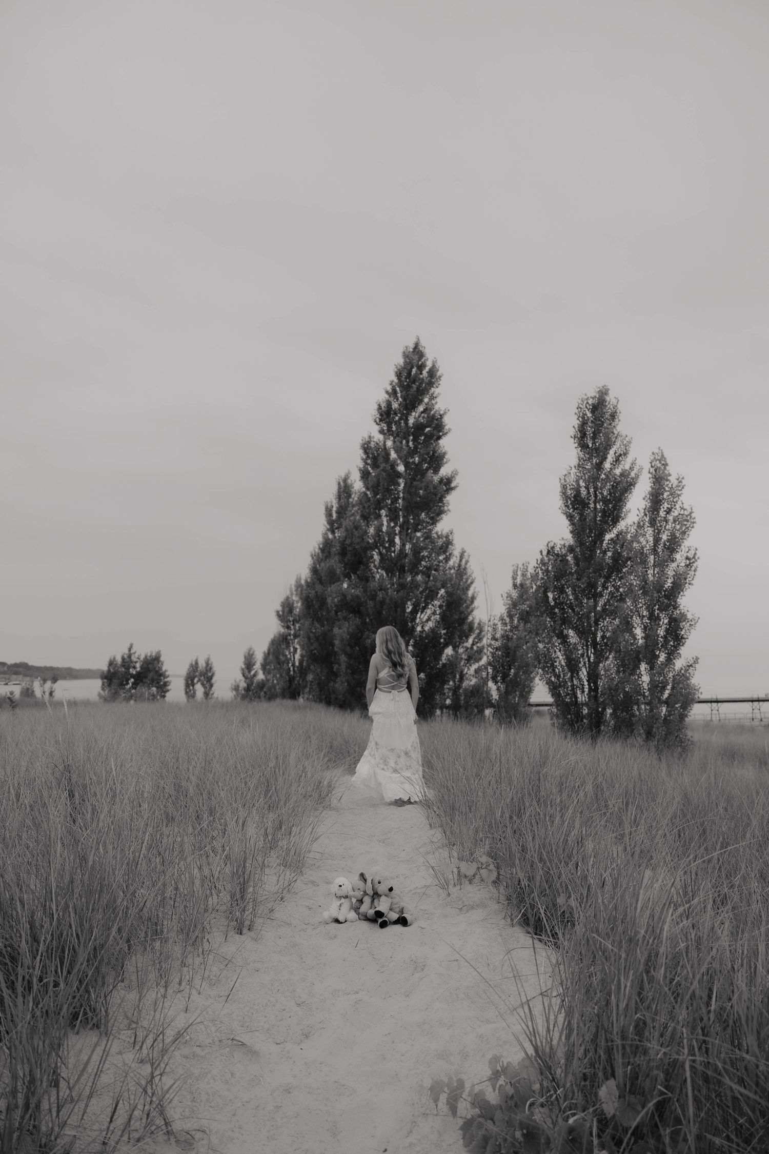 Black and white photo of a bride in a white gown walking a sandy path toward tall trees in an open field.