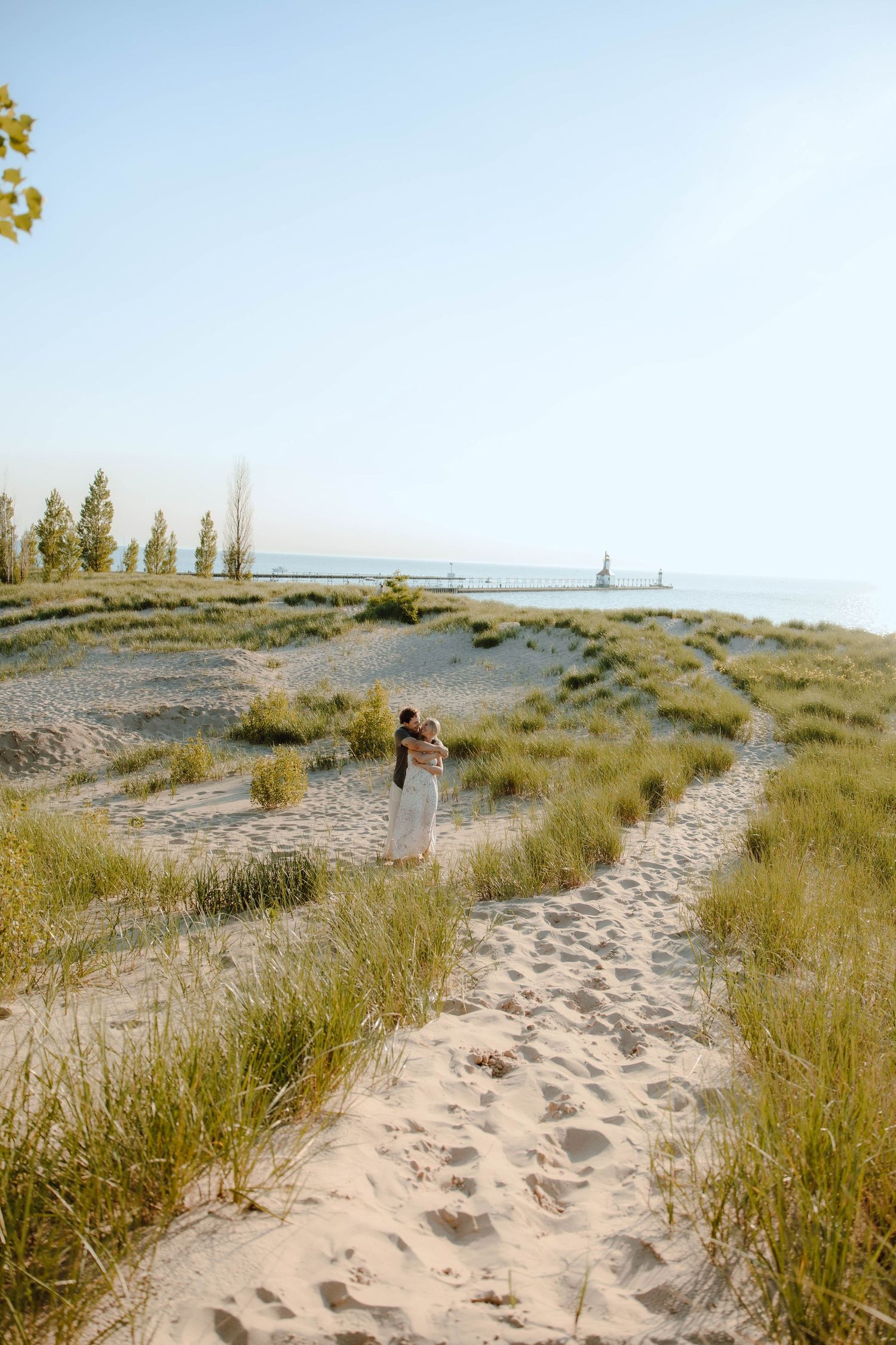 Couple embracing on a sandy beach path surrounded by dune grass with a lighthouse visible in the distance.