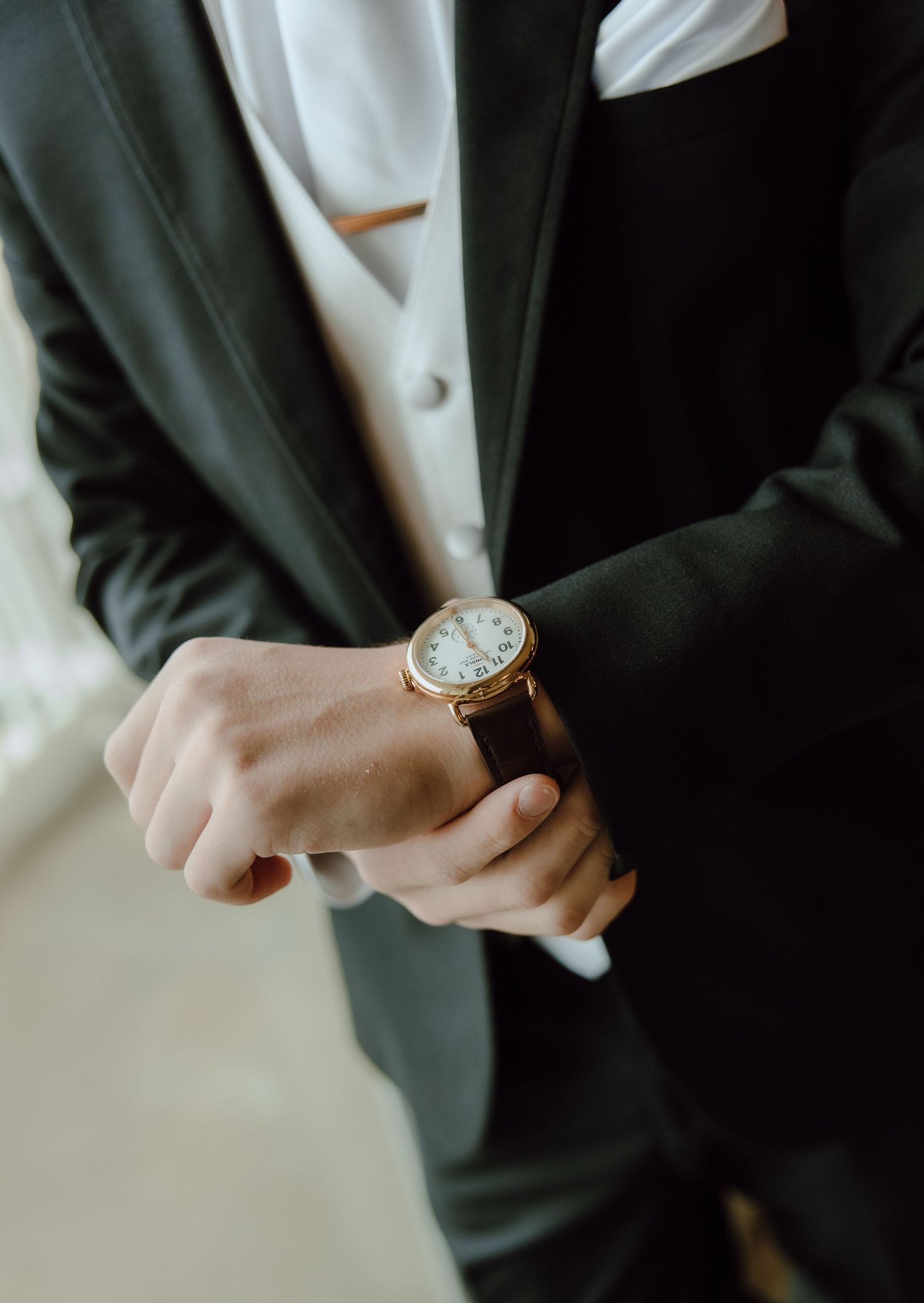 Groom in black suit adjusting gold watch on wrist before wedding ceremony.