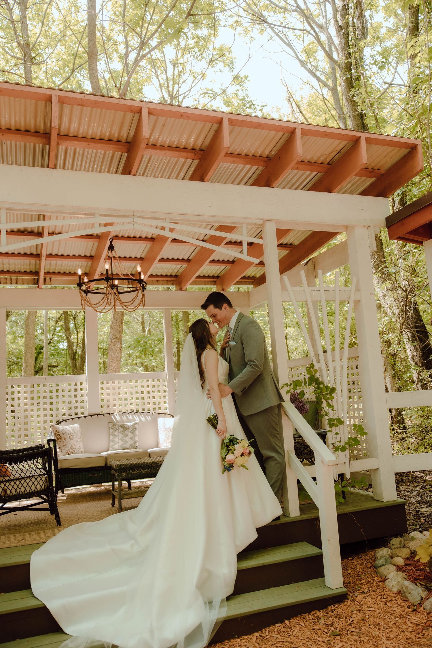 Bride and groom share a romantic kiss under a white pergola surrounded by lush green trees at an outdoor wedding.