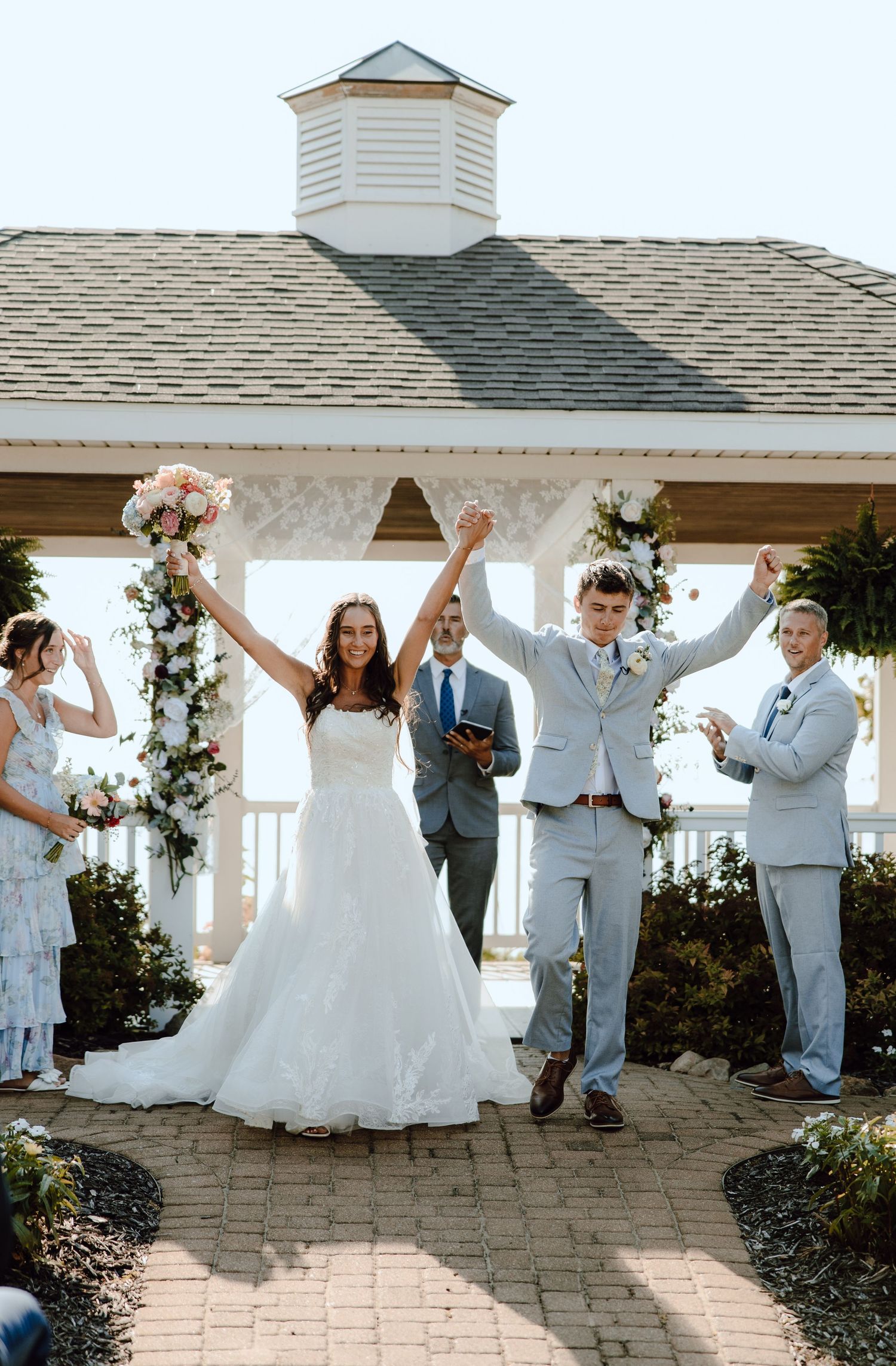 Bride and groom celebrating with raised hands at gazebo wedding ceremony under blue sky.