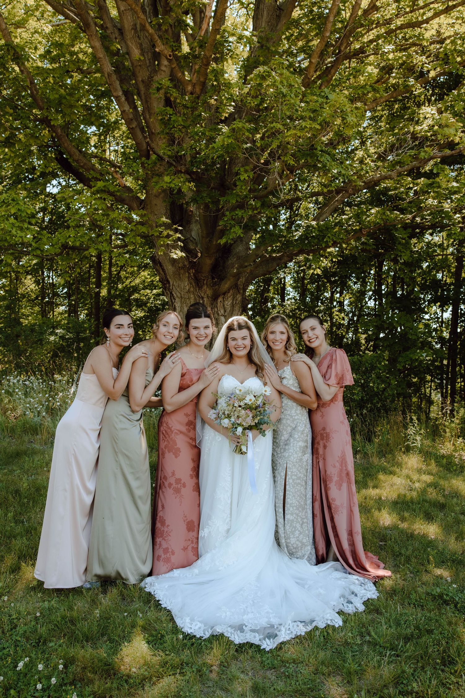 Smiling bride in white lace gown poses with five bridesmaids in mixed earth-tone dresses under a large tree.