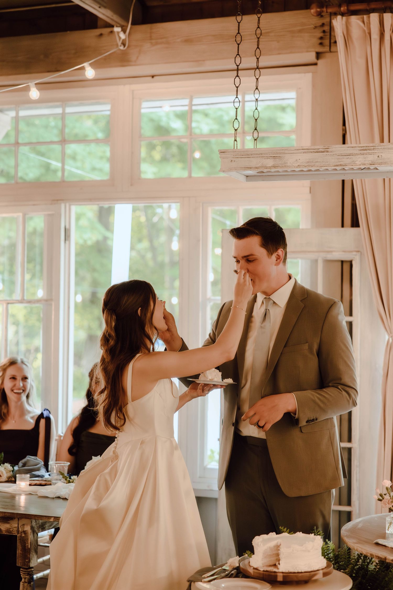 Bride feeds wedding cake to groom during reception as guests smile in a warmly lit rustic venue.