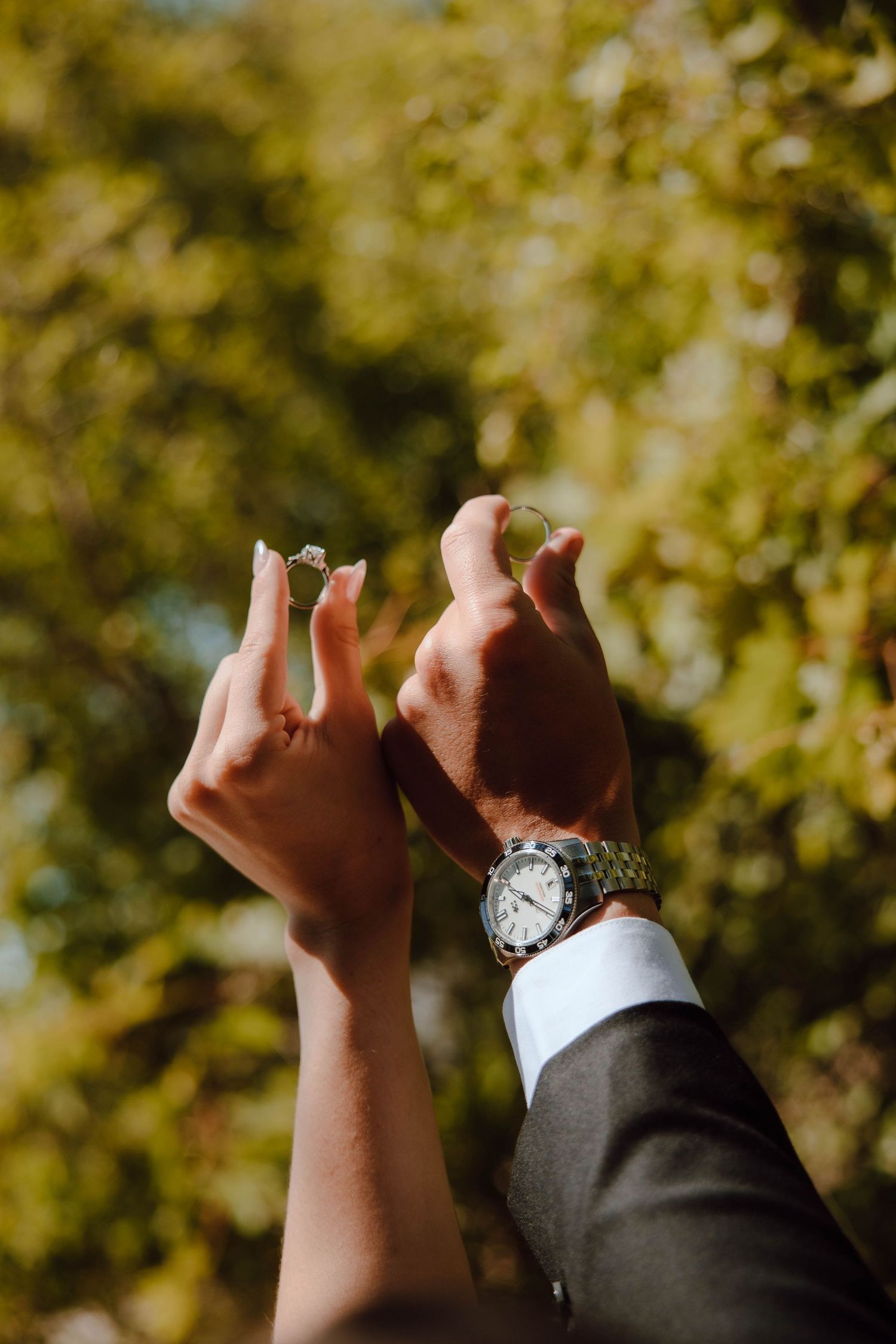 Bride and groom raising hands with wedding rings outdoors, with lush green trees in the background on a sunny day.