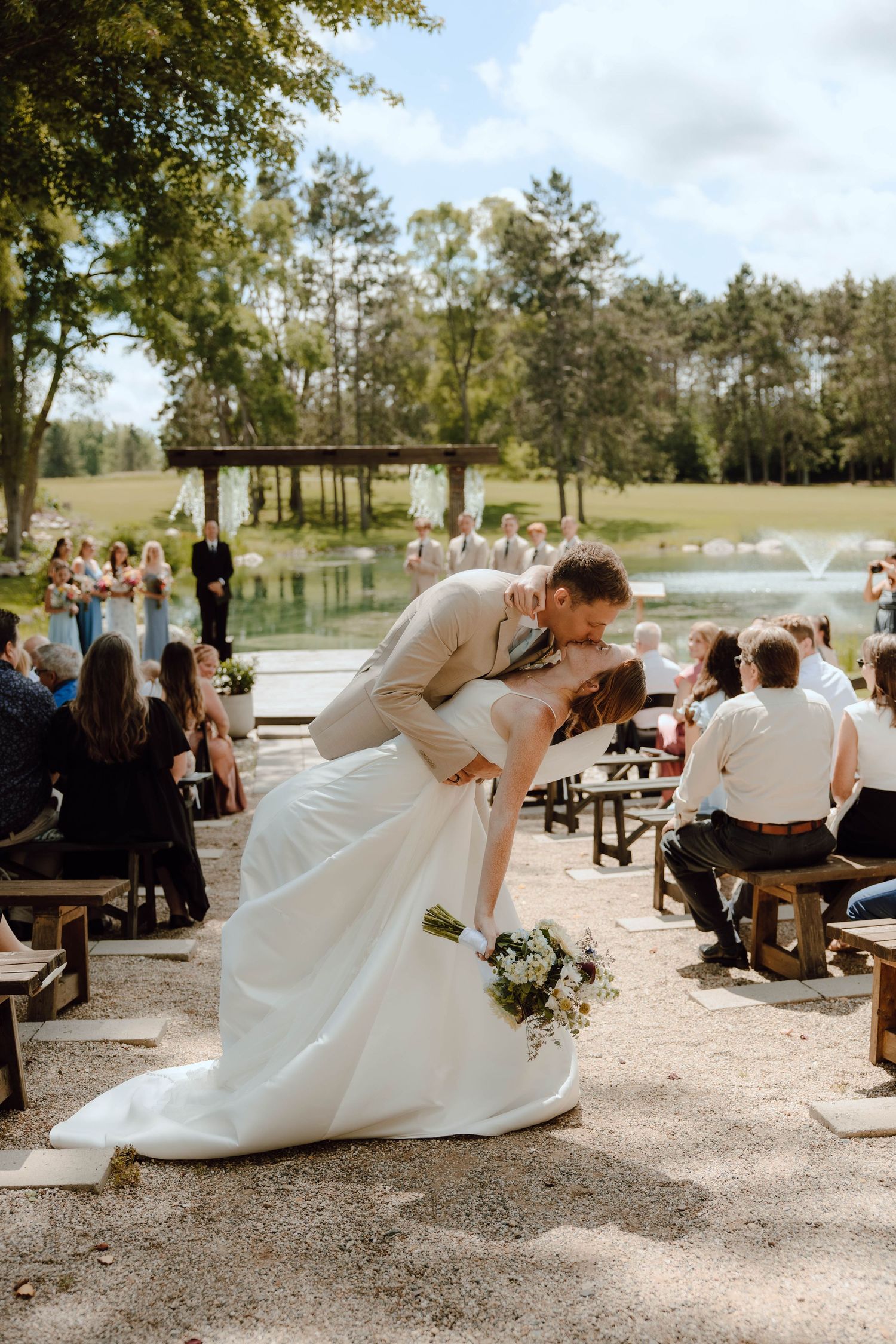 Bride and groom share a romantic dip kiss at outdoor wedding ceremony near a pond surrounded by seated guests.