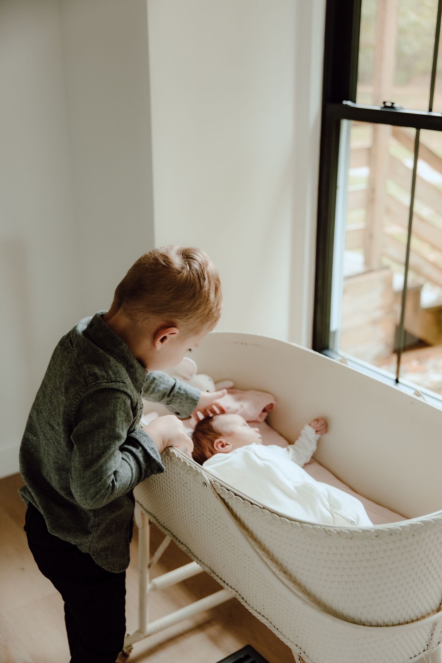 Child looking into white bassinet near window in modern nursery setting.