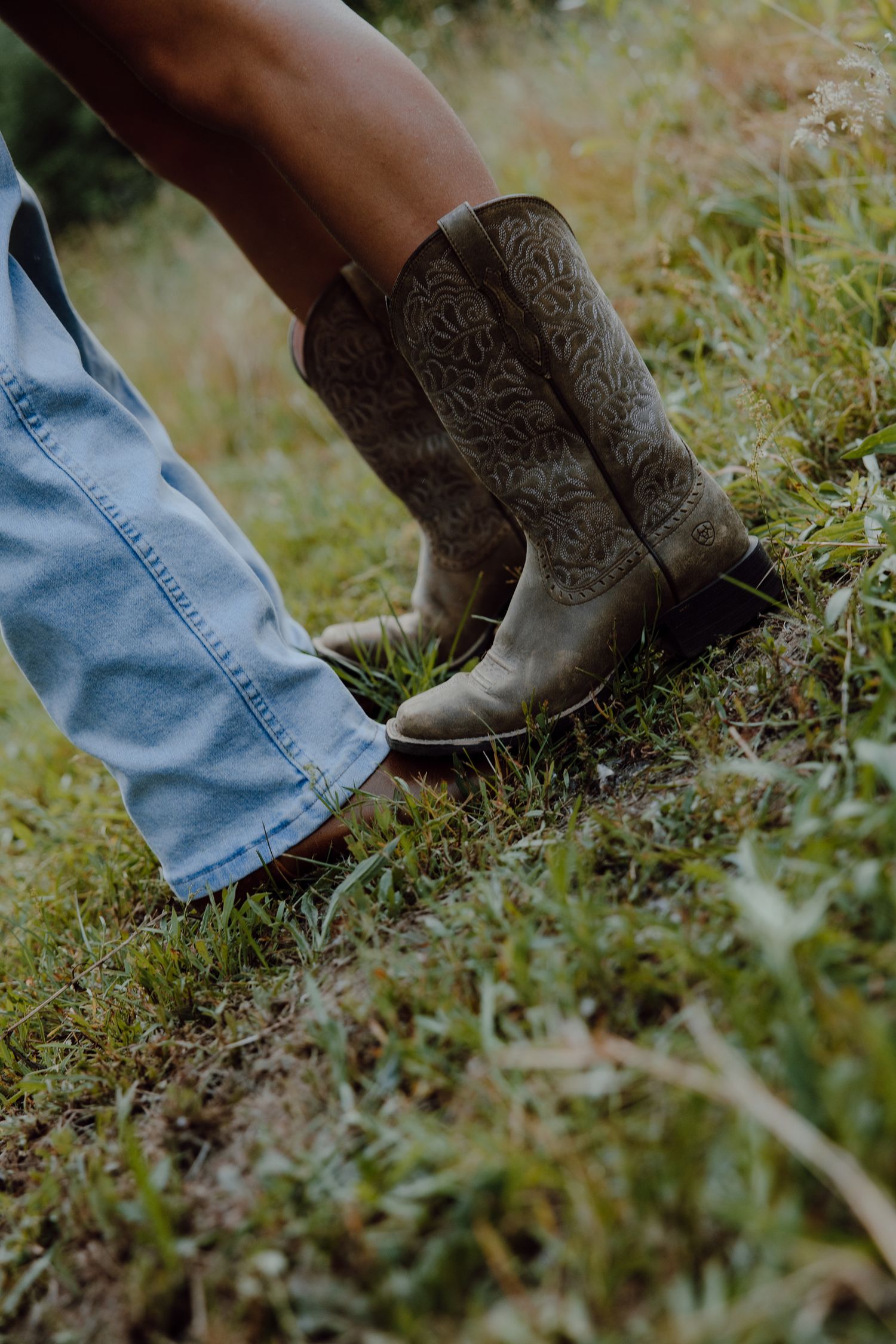 Brown leather cowboy boots with intricate embroidery detail standing in grass and wildflowers.