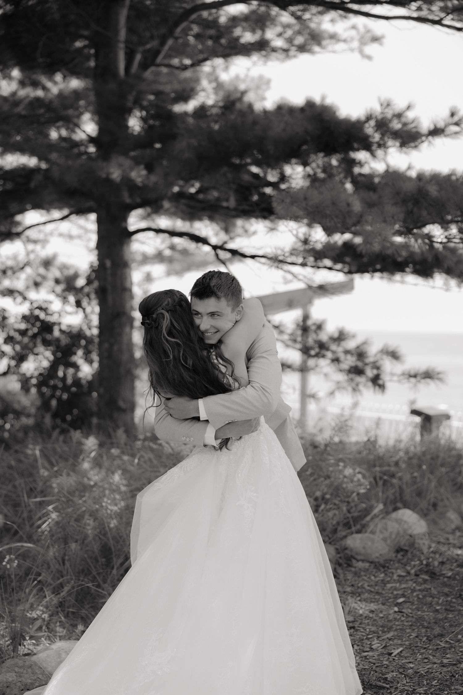 A bride and groom share a romantic embrace among pine trees with a scenic waterfront backdrop.