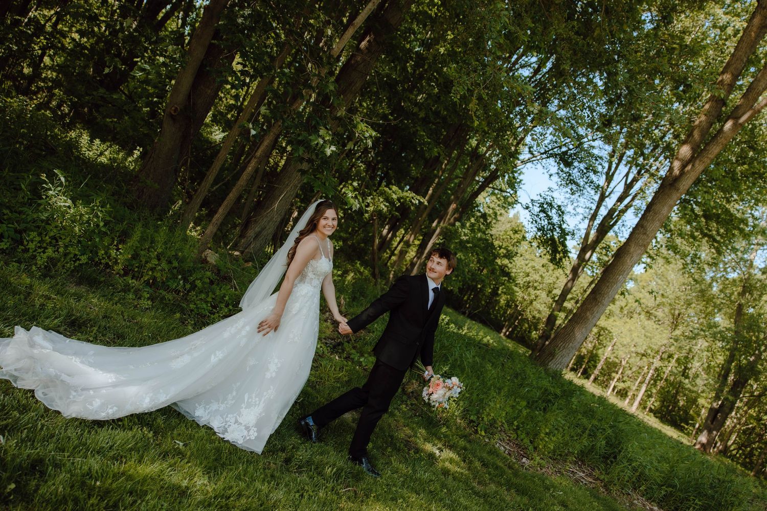 Bride in white gown and groom in black suit walk hand-in-hand through a lush green forest on their wedding day.