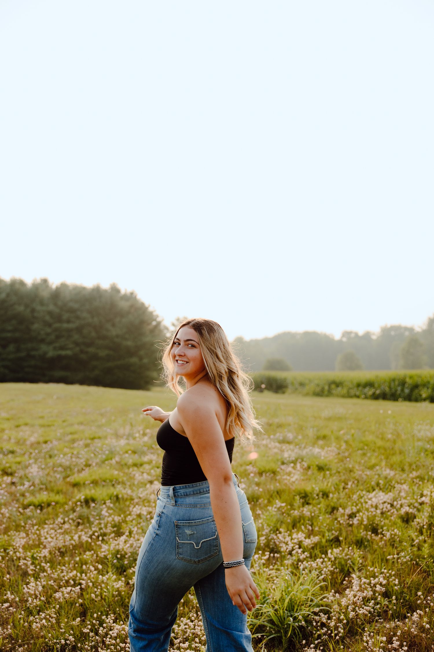 Person in black top and jeans poses in sunlit meadow with forest backdrop.