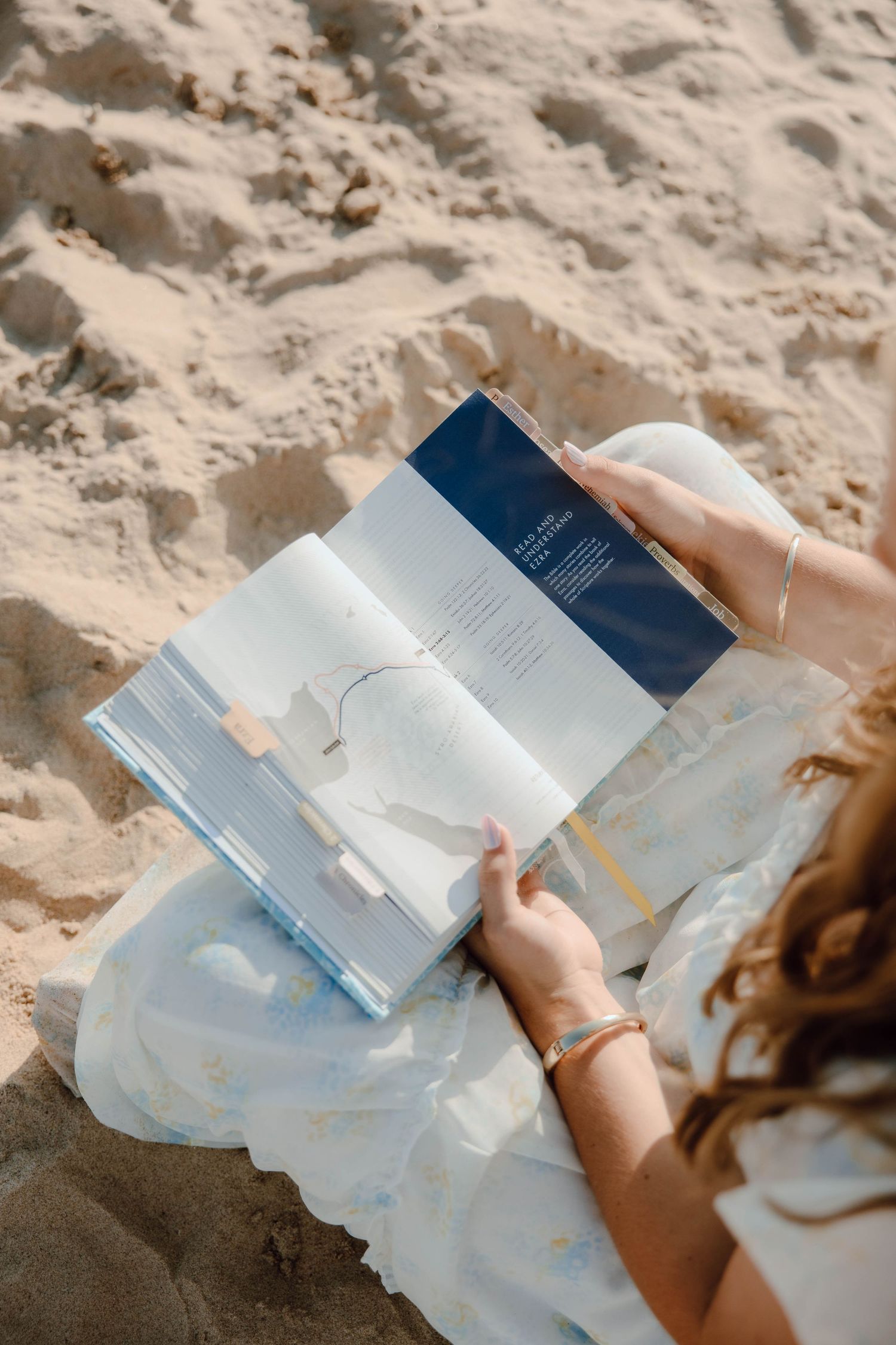 Woman reading magazine while relaxing on sandy beach in white dress.