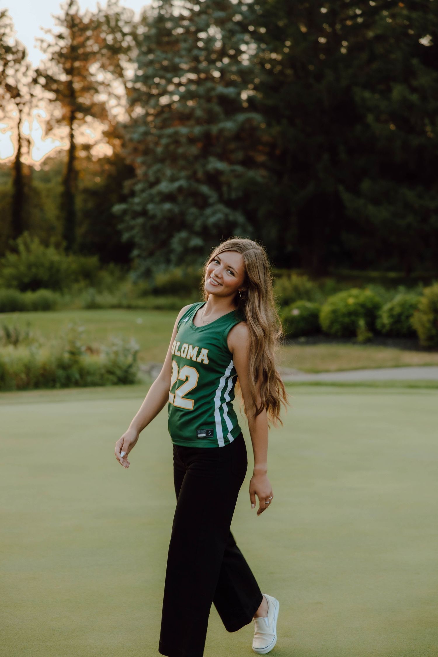 Smiling teenage girl in green sports jersey number 22 walking on golf course at sunset.