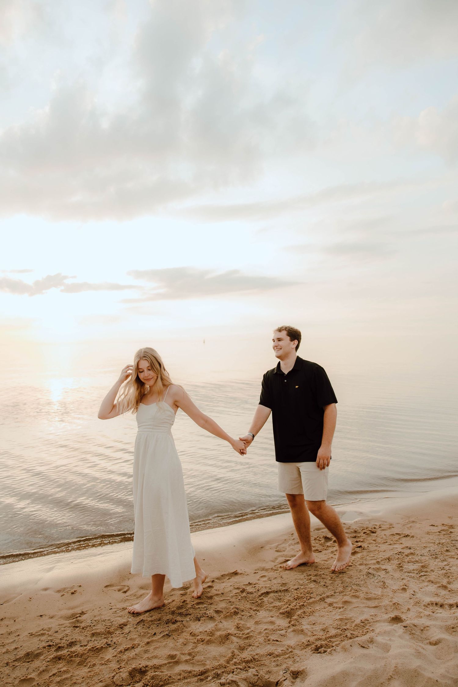 Couple holding hands walking barefoot on sandy beach at sunset, woman in white dress, man in black shirt.