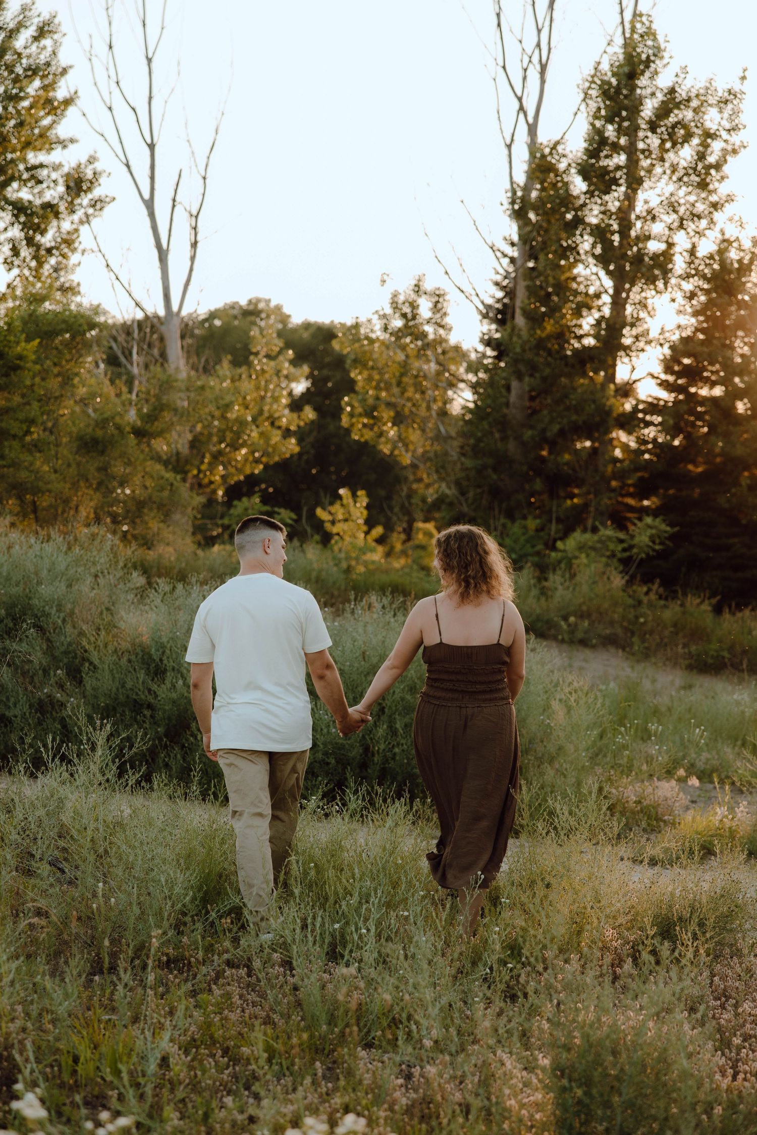 Couple holding hands walking through a lush green meadow at golden hour sunset.