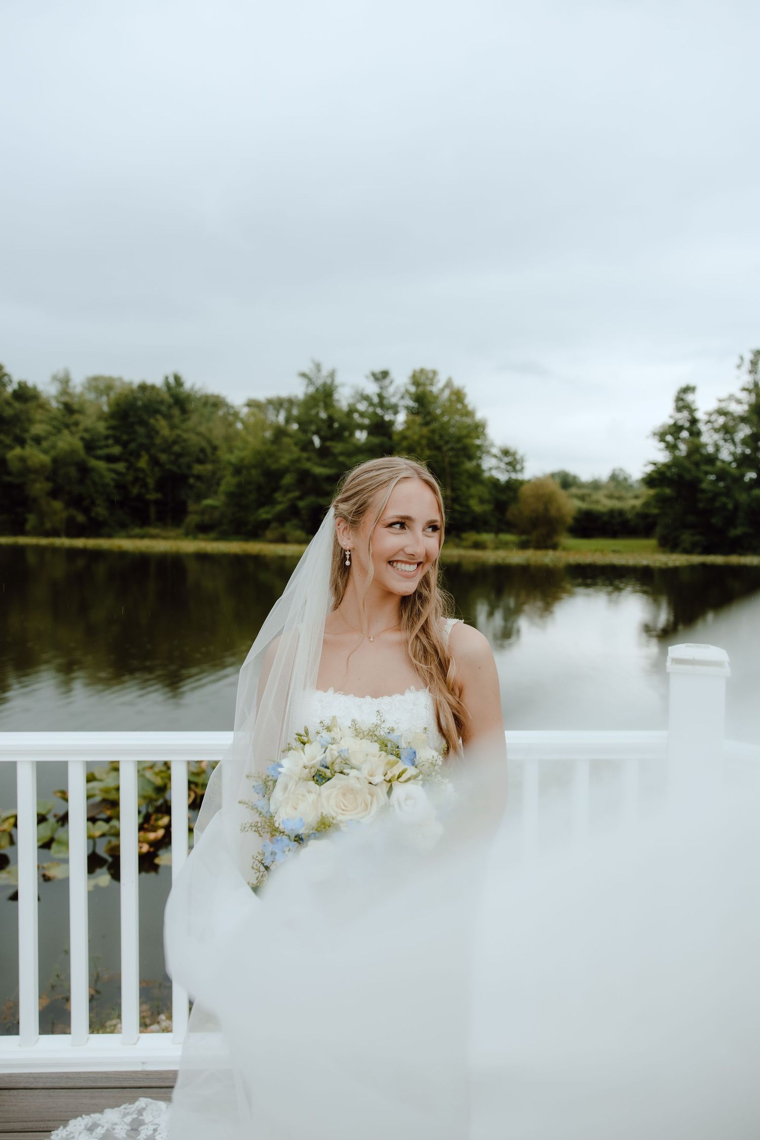 Radiant bride holding white bouquet poses on white deck overlooking serene pond.