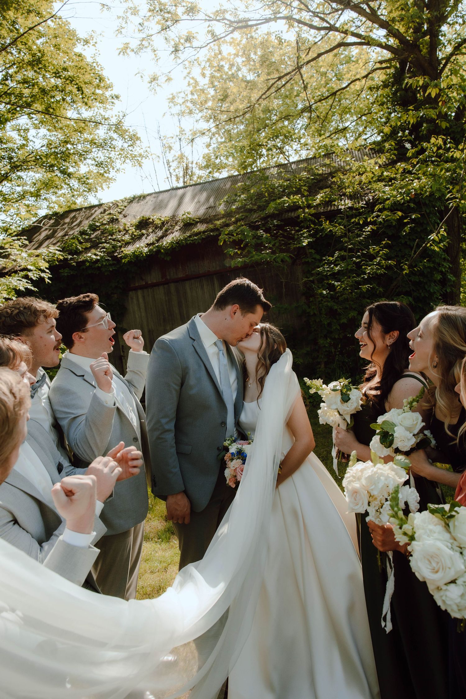 Newlyweds kiss surrounded by smiling wedding party holding white bouquets in sunny outdoor setting.